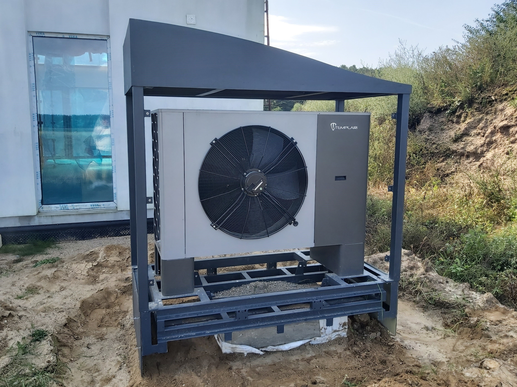 Zewnętrzna pompa ciepła Templari, umieszczona na metalowej konstrukcji z zadaszeniem, w pobliżu budynku z oknem, na tle zieleni. Montaż w trakcie prac ziemnych.
