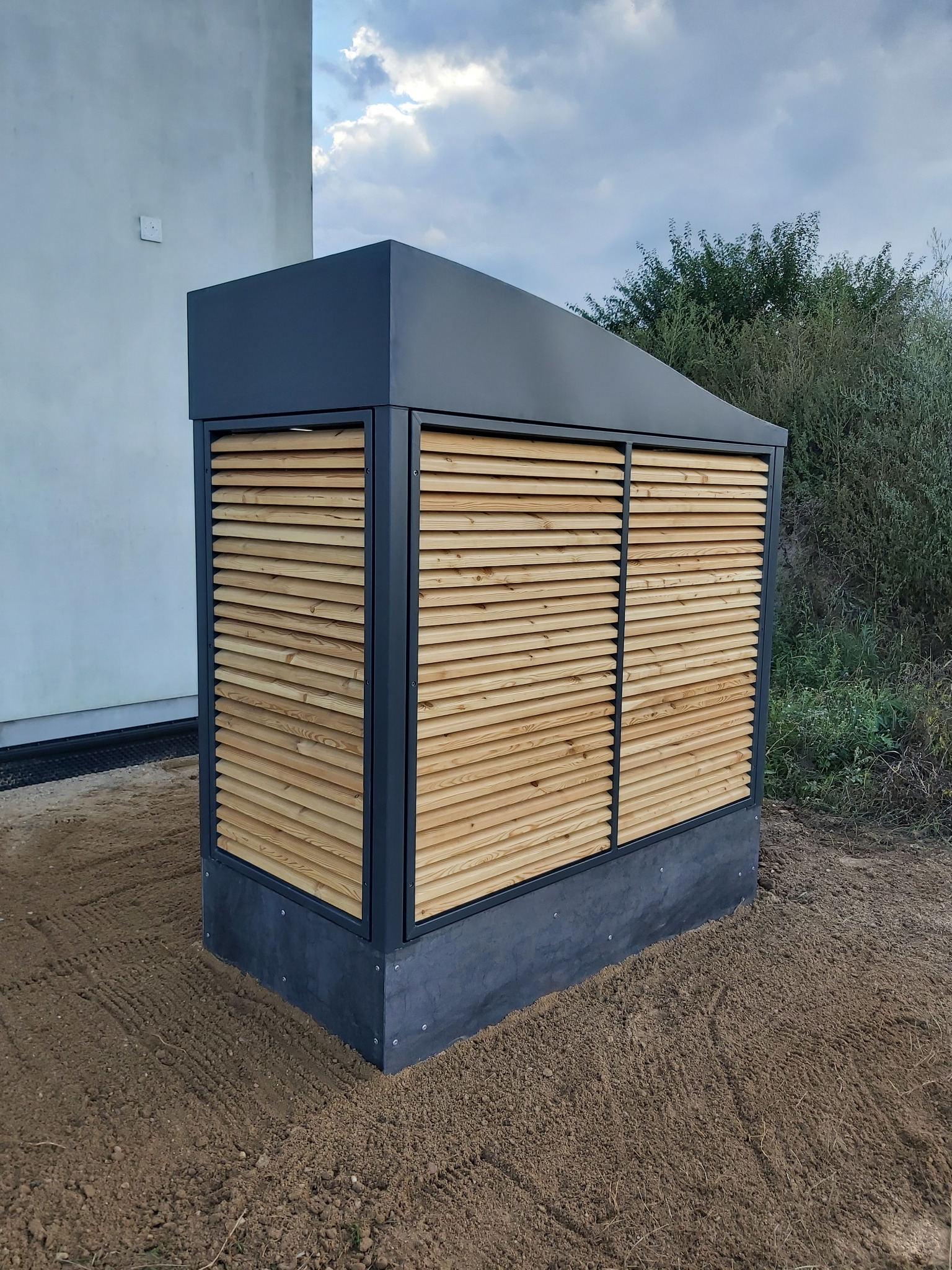Zewnętrzna obudowa jednostki wentylacyjnej z drewnianymi żaluzjami, umieszczona na ziemi przy budynku. Szara obudowa, jasne drewno, widoczne tło z zielenią.