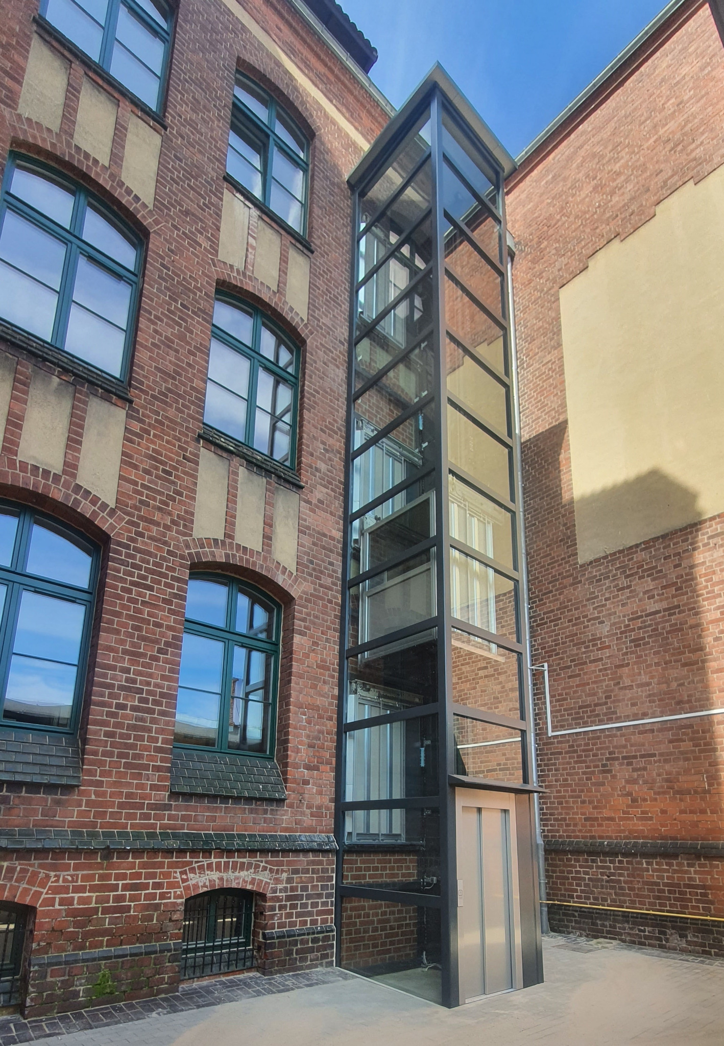 Zewnętrzna winda osobowa z przeszkloną konstrukcją zamontowana przy budynku z cegły w Zielonej Górze. Widoczne okna z zielonymi ramami i fragment nieba.