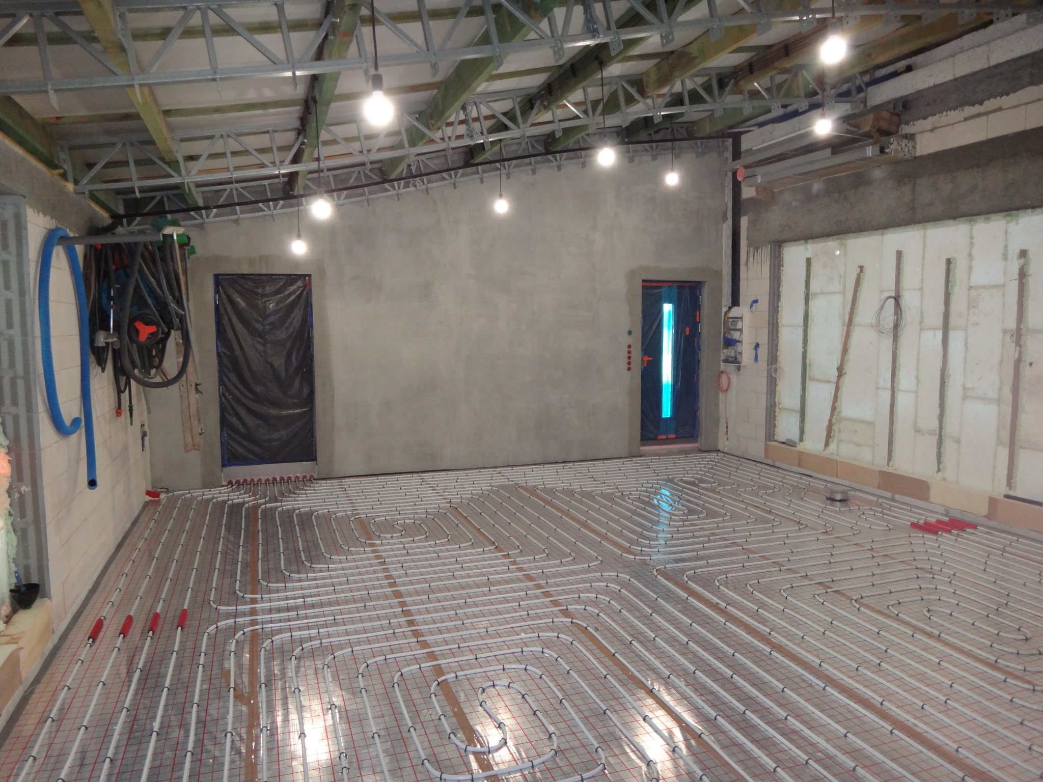 Rozłożona instalacja ogrzewania podłogowego na siatce w surowym wnętrzu. Widoczne białe rury, czerwone złączki, surowy beton i elementy konstrukcji sufitu.