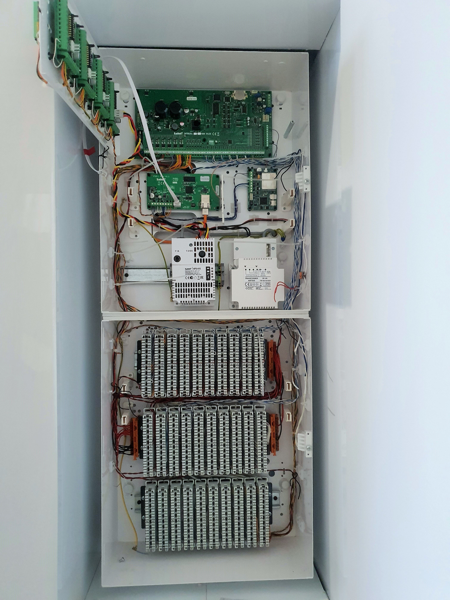 Otwarta skrzynka elektryczna z licznymi przewodami, płytkami drukowanymi i złączkami, zamontowana na białej ścianie. Widok z góry.