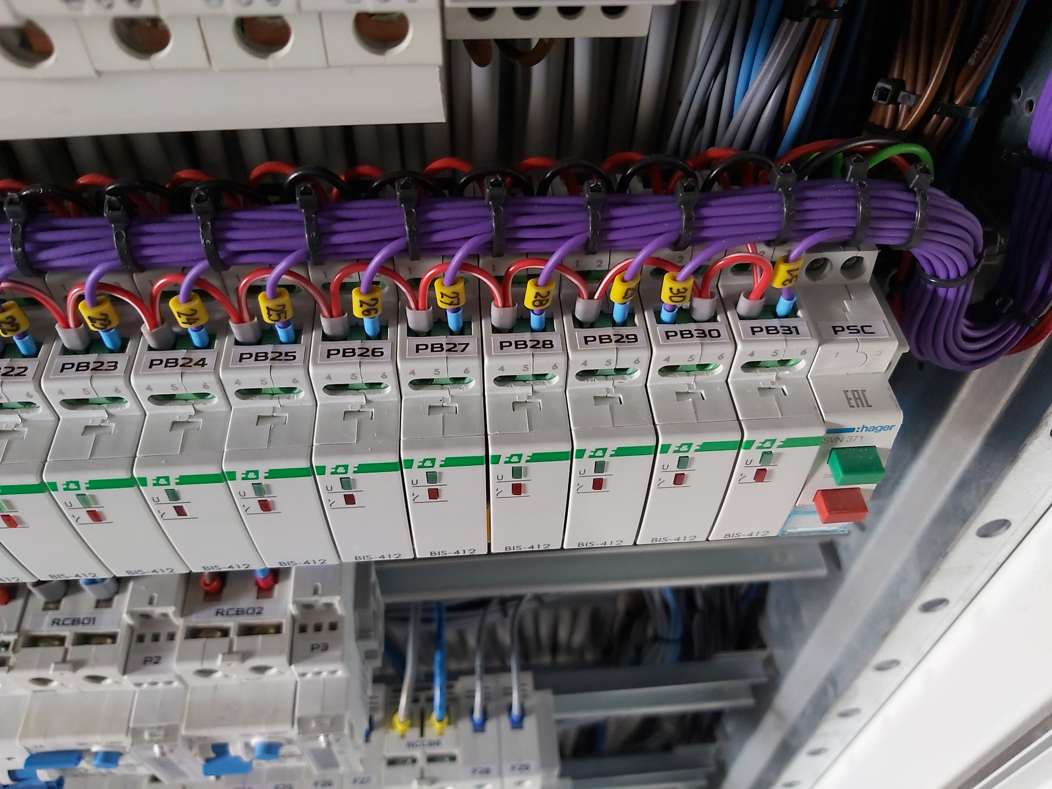 Szczegółowe ujęcie rozdzielnicy elektrycznej z oznaczonymi przewodami i bezpiecznikami. Widoczne kable w kolorach fioletowym, czerwonym i niebieskim.