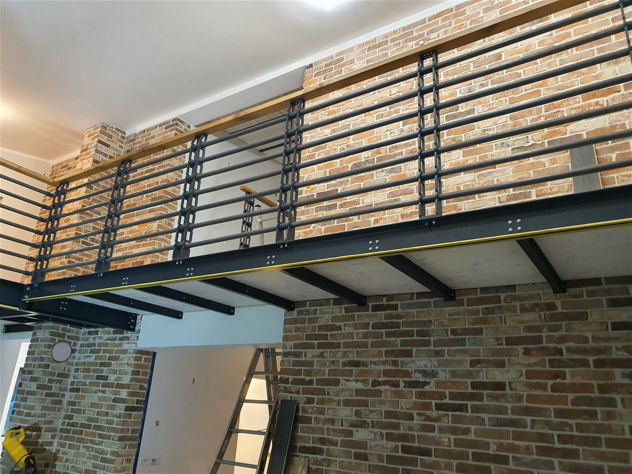 Wnętrze z antresolą wykończoną cegłą i metalową balustradą, widok z dołu. Widoczna drabina i narzędzia, sugerujące trwające prace wykończeniowe.