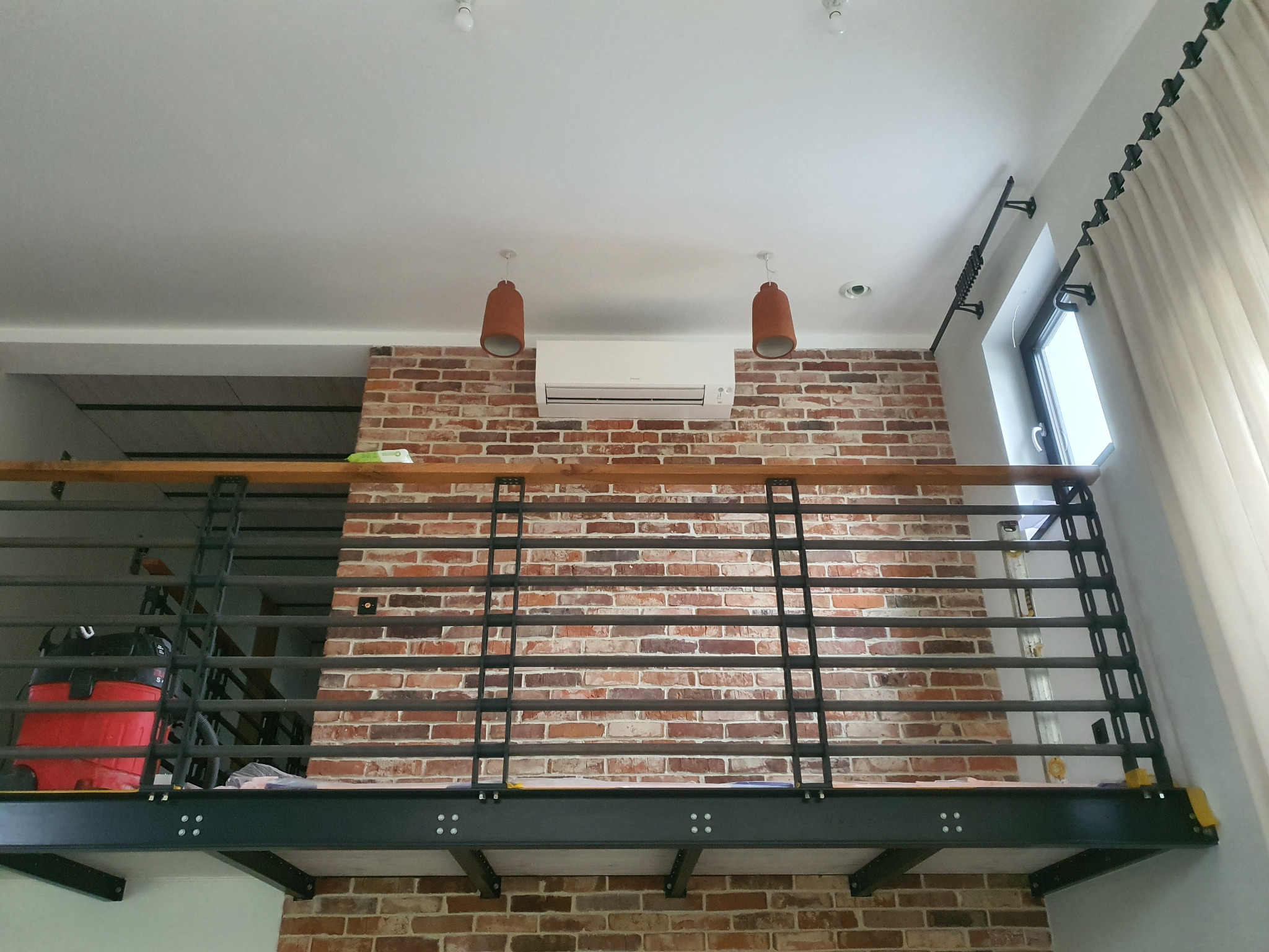 Loft z klimatyzacją na ceglanej ścianie, widok z dołu. Metalowa balustrada, wiszące lampy i okno z zasłoną. Czerwony odkurzacz na antresoli.