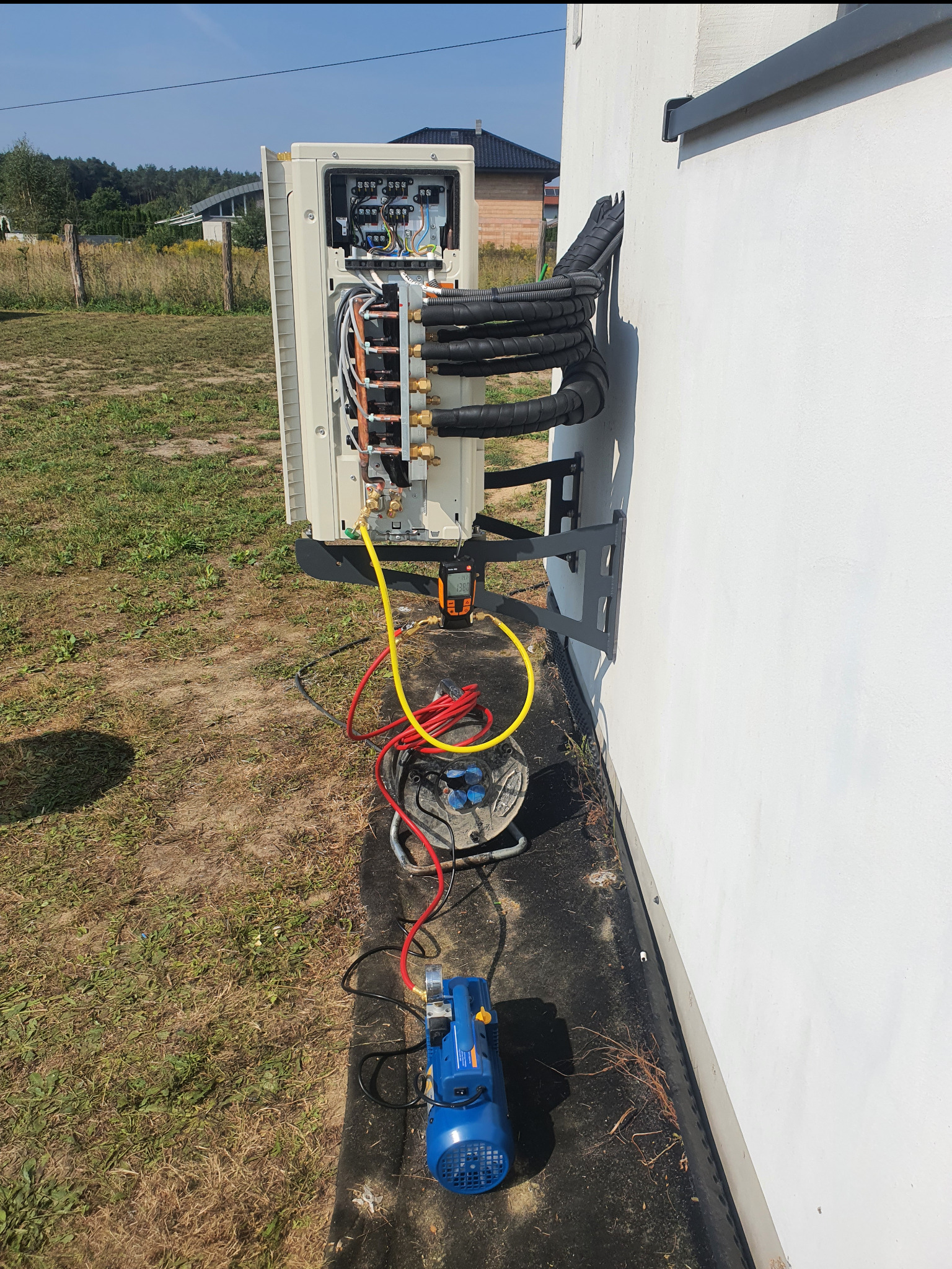 Montaż klimatyzacji zewnętrznej na białej ścianie budynku. Widoczne przewody, narzędzia i elementy instalacji. Trawa i drzewa w tle. Dzień słoneczny.