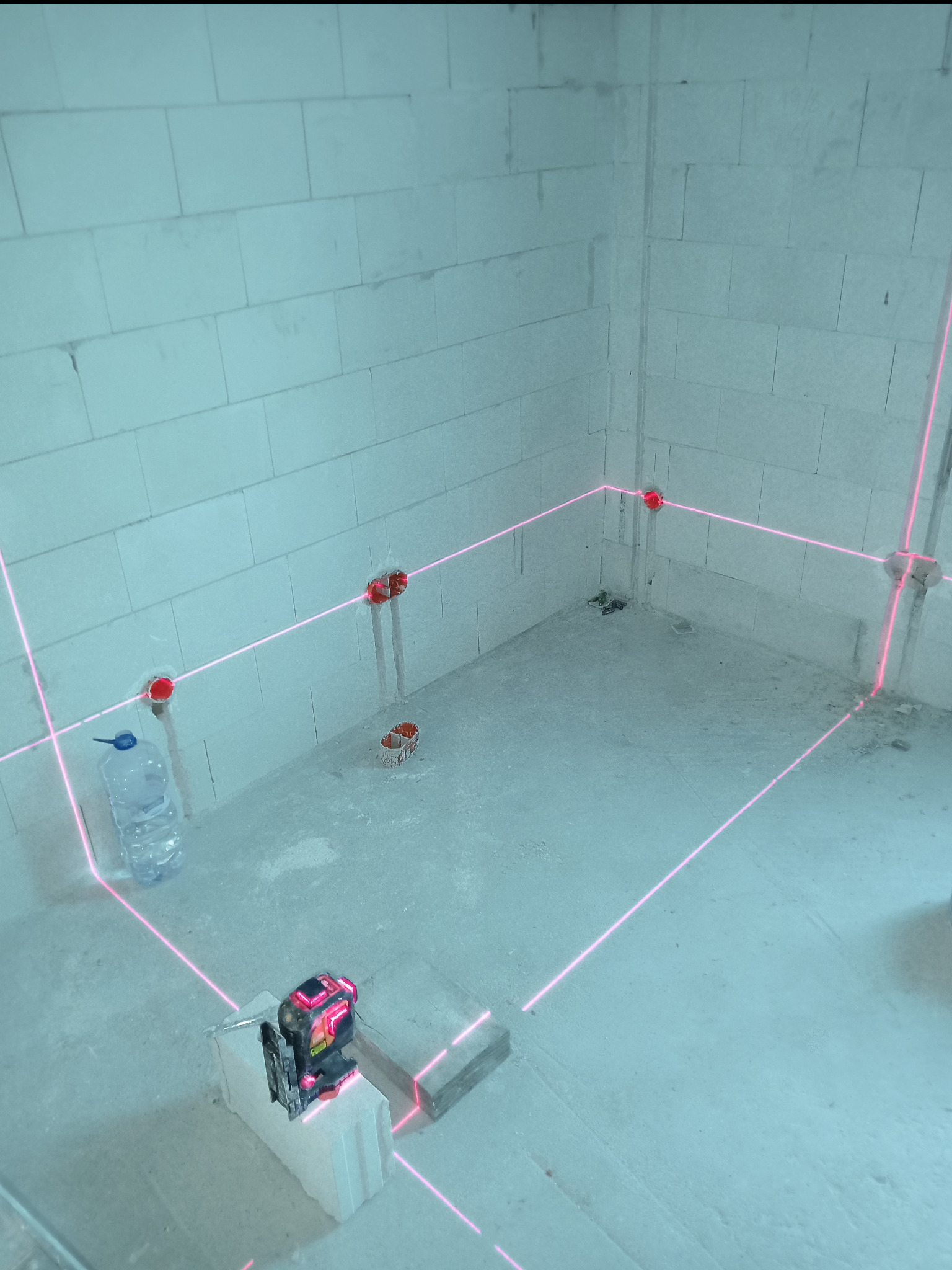 Surowy pokój z betonu komórkowego z wyznaczonym laserowo poziomem instalacji elektrycznej. Widoczne puszki elektryczne i butelka wody.