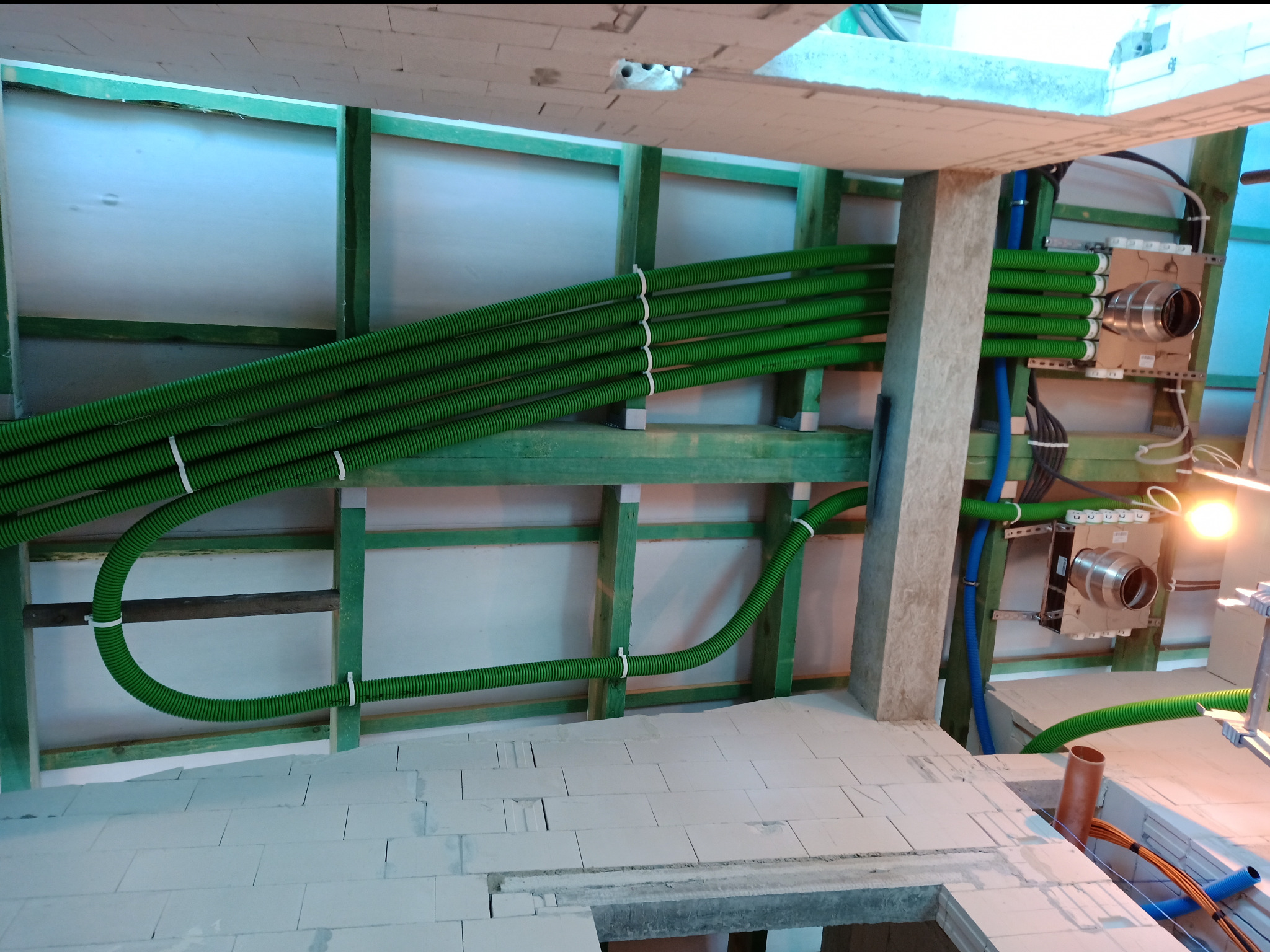 Wnętrze w trakcie instalacji: zielone rury wentylacyjne przymocowane do drewnianej konstrukcji. Widoczne również niebieskie rury i fragment podłogi z białych bloczków.