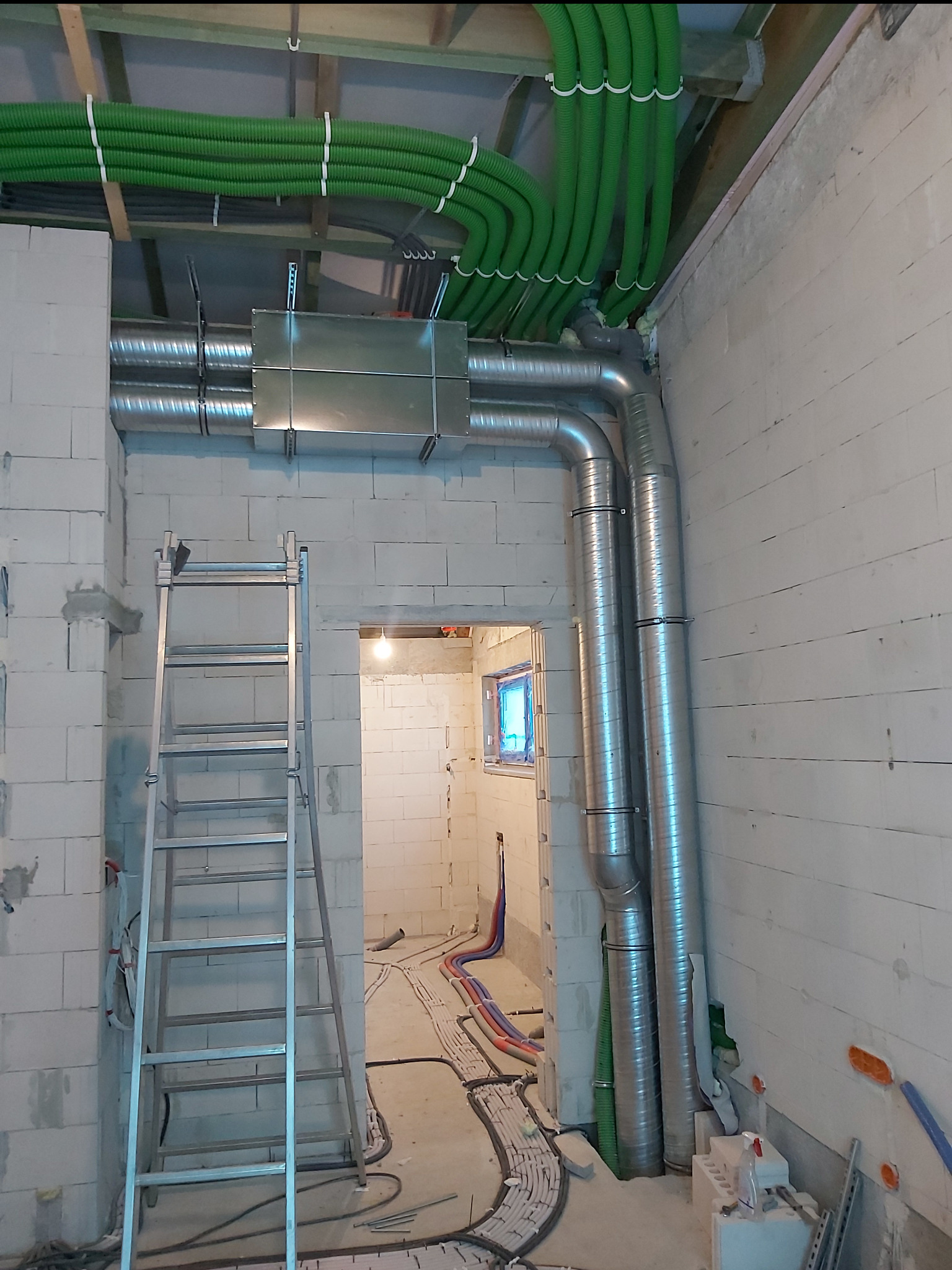 Widok na sufit z zielonymi rurami wentylacyjnymi, srebrne rury pionowe i rozgałęzienia. W tle widoczna drabina i instalacje podłogowe w surowym wnętrzu.