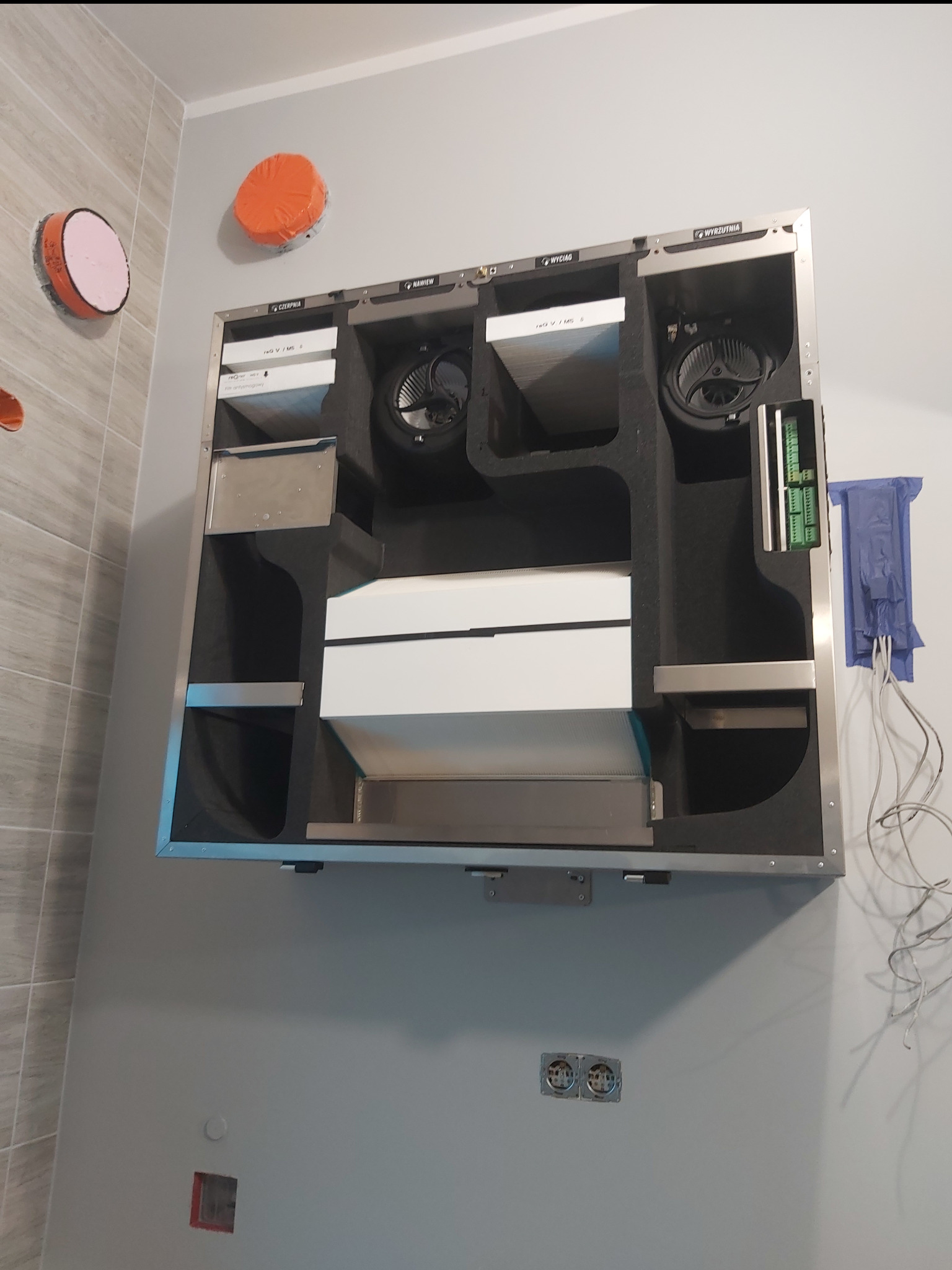 Centrala wentylacyjna z widocznymi filtrami i wentylatorami zamontowana na ścianie w pomieszczeniu z elementami instalacji elektrycznej.