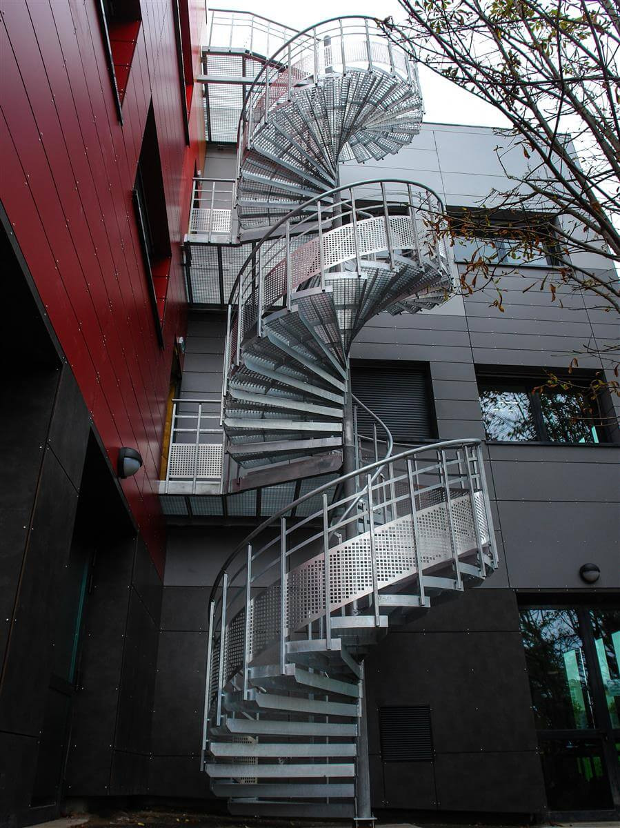 Zewnętrzne metalowe schody spiralne przy budynku o czerwono-szarej elewacji. Widok z dołu, ukazujący konstrukcję i detale schodów.
