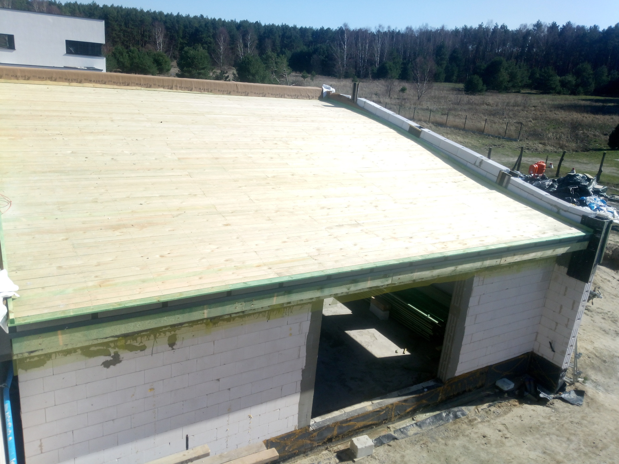 Budowa domu: białe bloczki, drewniana konstrukcja dachu z deskowaniem, widok z góry na plac budowy i las w tle.