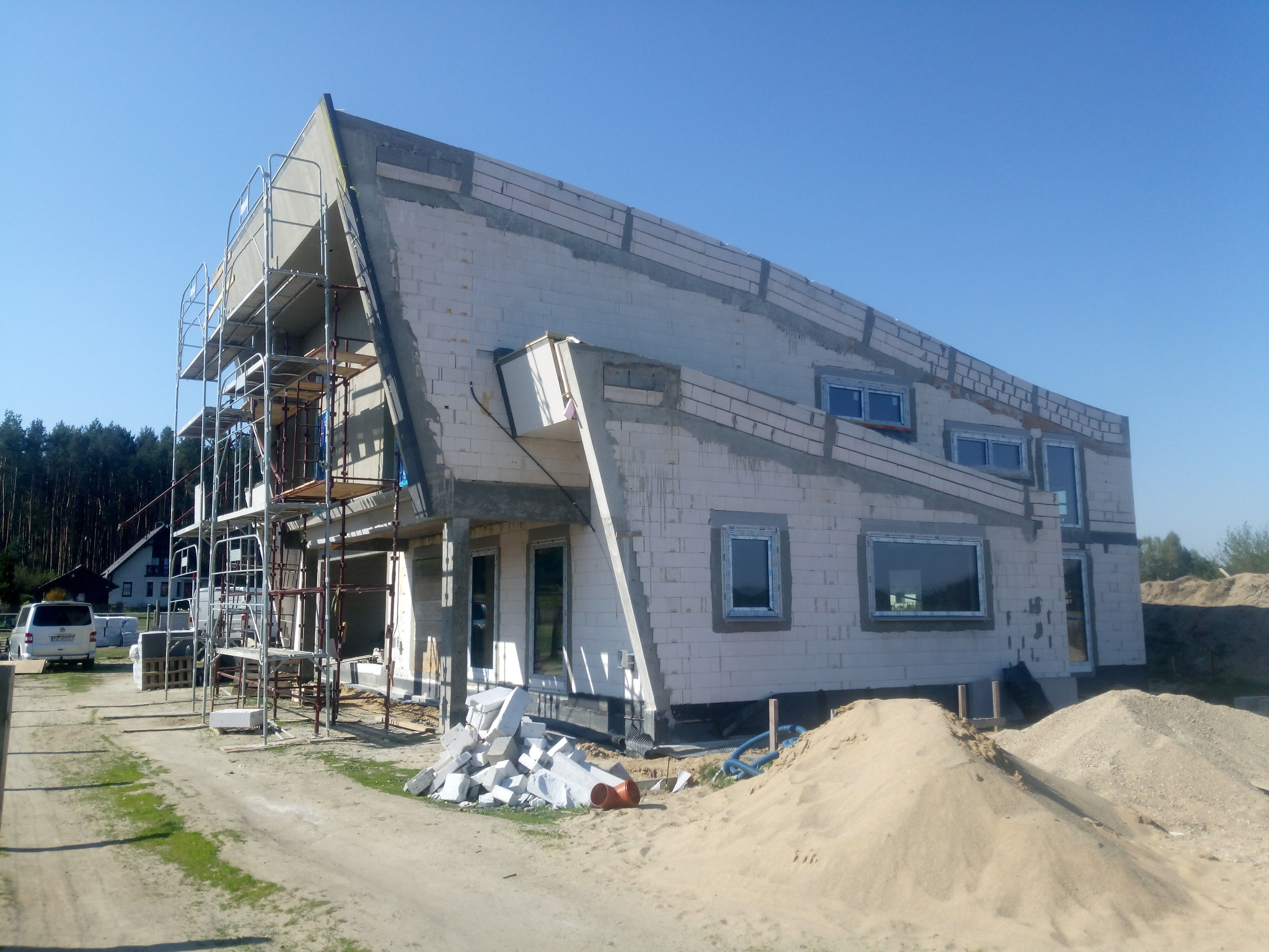 Surowy stan domu z białego muru, w trakcie budowy, z rusztowaniami i stertą piasku. Widoczne okna i konstrukcja dachu. Błękitne niebo w tle.