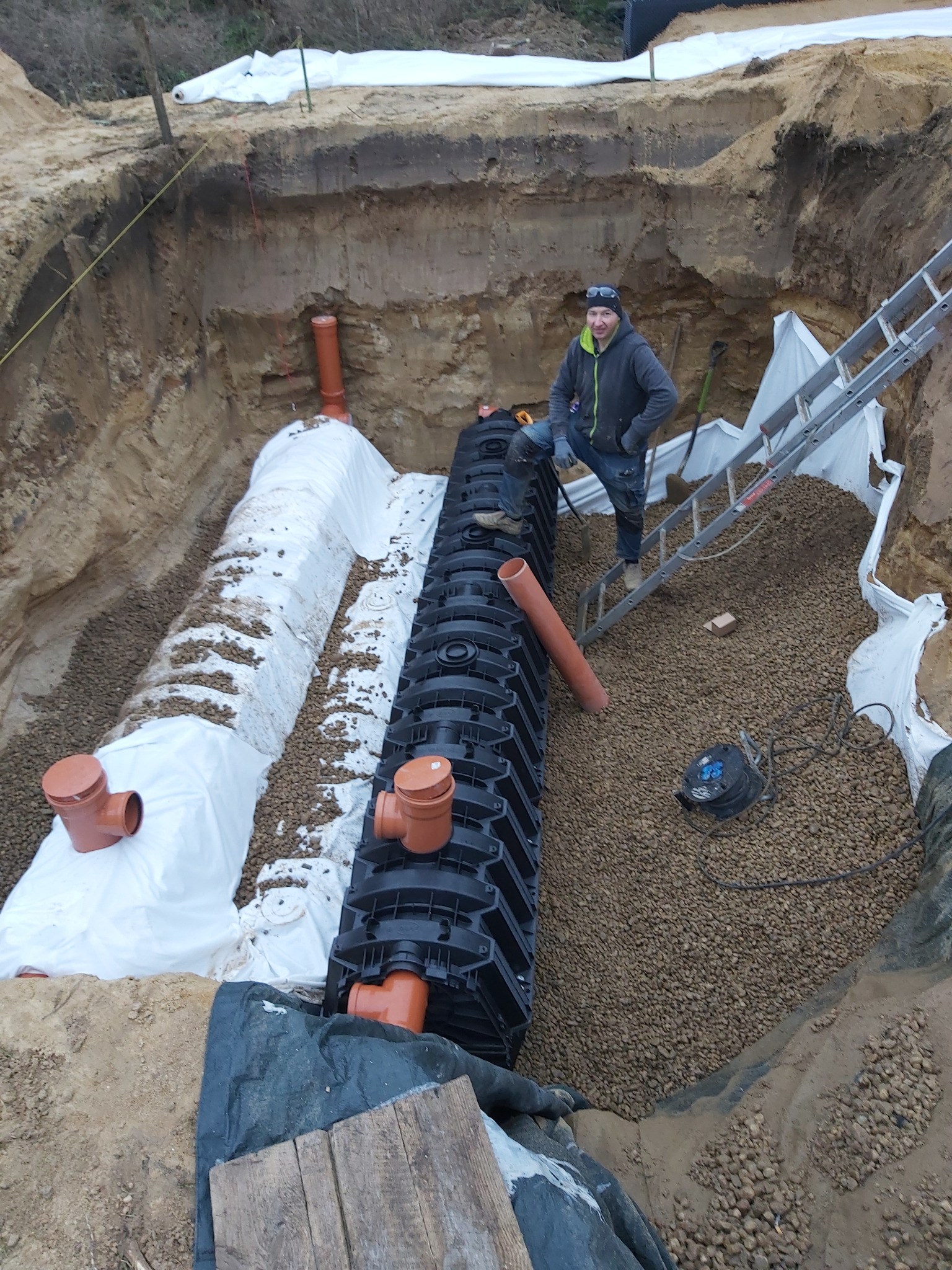 Instalacja kanalizacji deszczowej w wykopie ziemnym. Czarna, modułowa konstrukcja, rury PCV, biała geowłóknina. Mężczyzna stoi na drabinie w tle.
