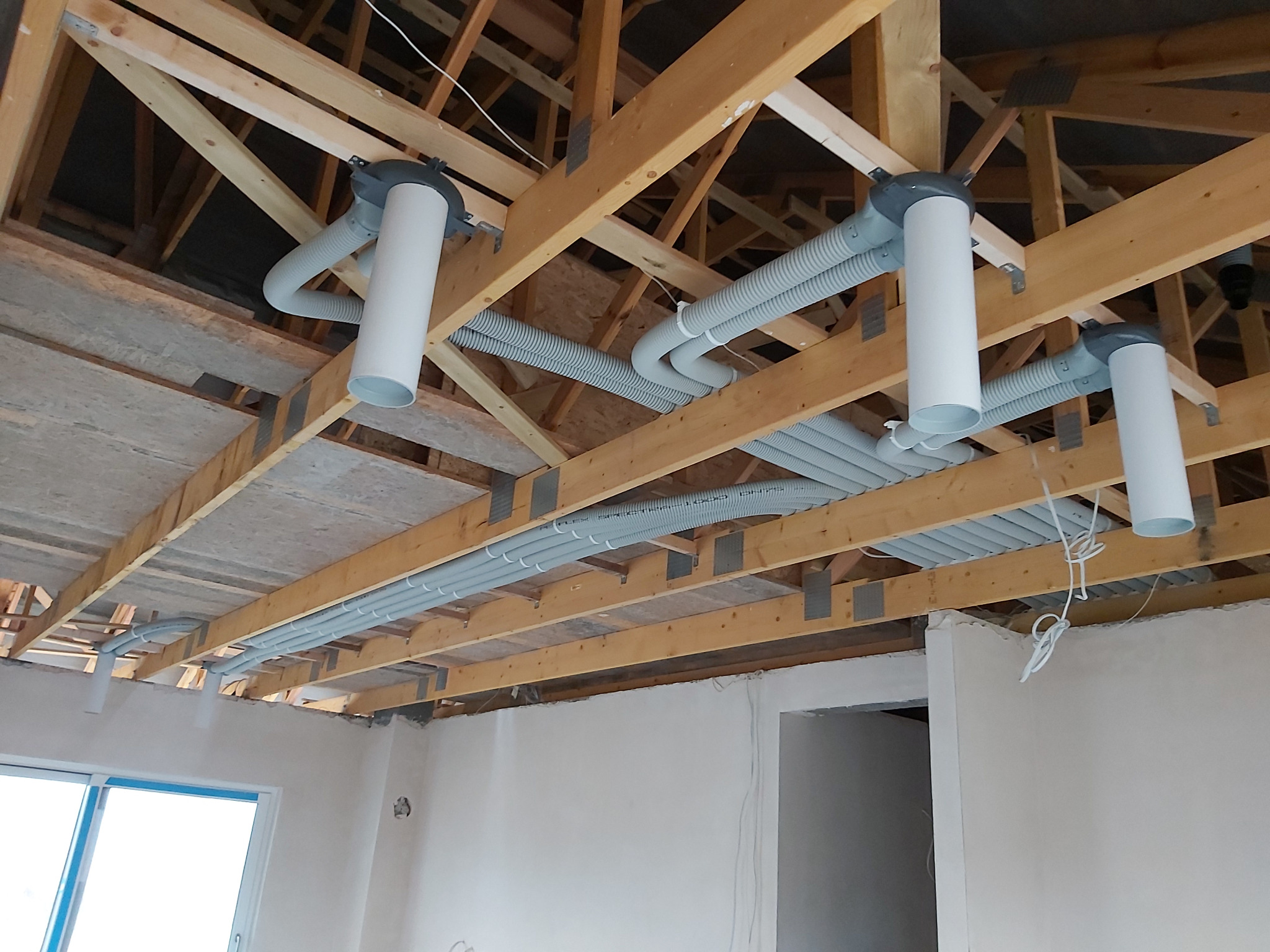 Wnętrze w trakcie montażu wentylacji mechanicznej: widoczne rury wentylacyjne pod stropem drewnianym, białe kanały wentylacyjne, surowy stan wykończenia ścian.