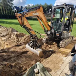 Żółto-czarna minikoparka Case w trakcie prac ziemnych na działce z trawą i kostką. Wykop pod instalacje lub fundamenty, widoczne drzewa i dom w tle.