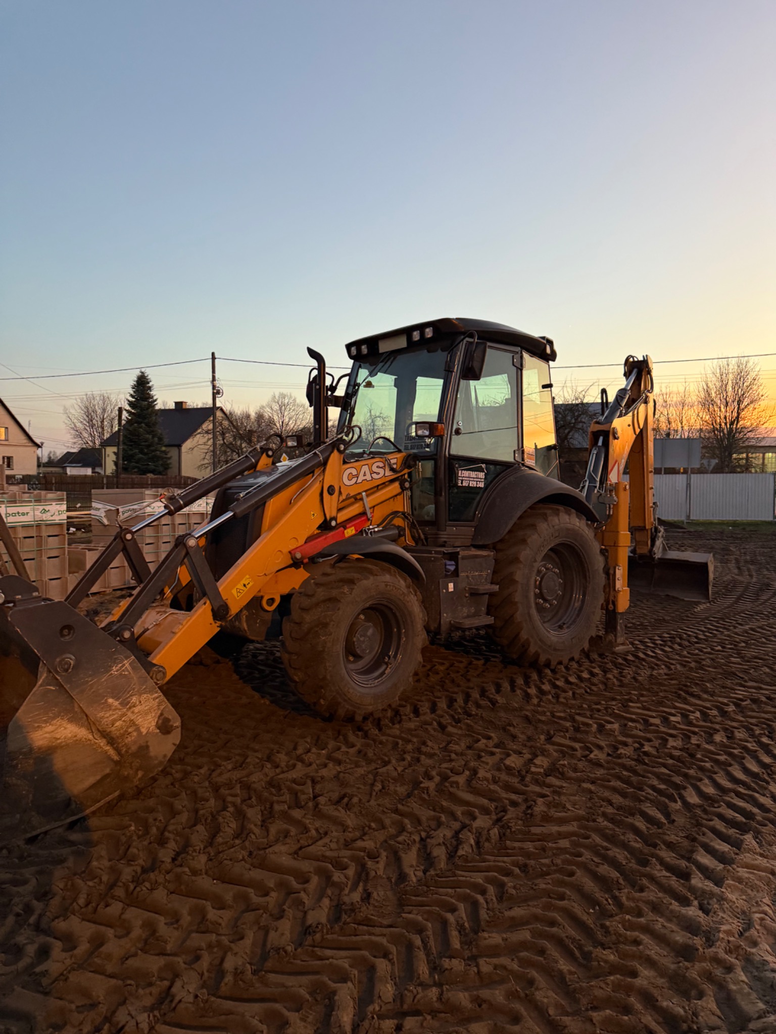 Żółto-czarna koparko-ładowarka Case na ujeżdżonym, błotnistym placu budowy o zmierzchu. Widoczny profil opony i ślady na ziemi. W tle budynki.