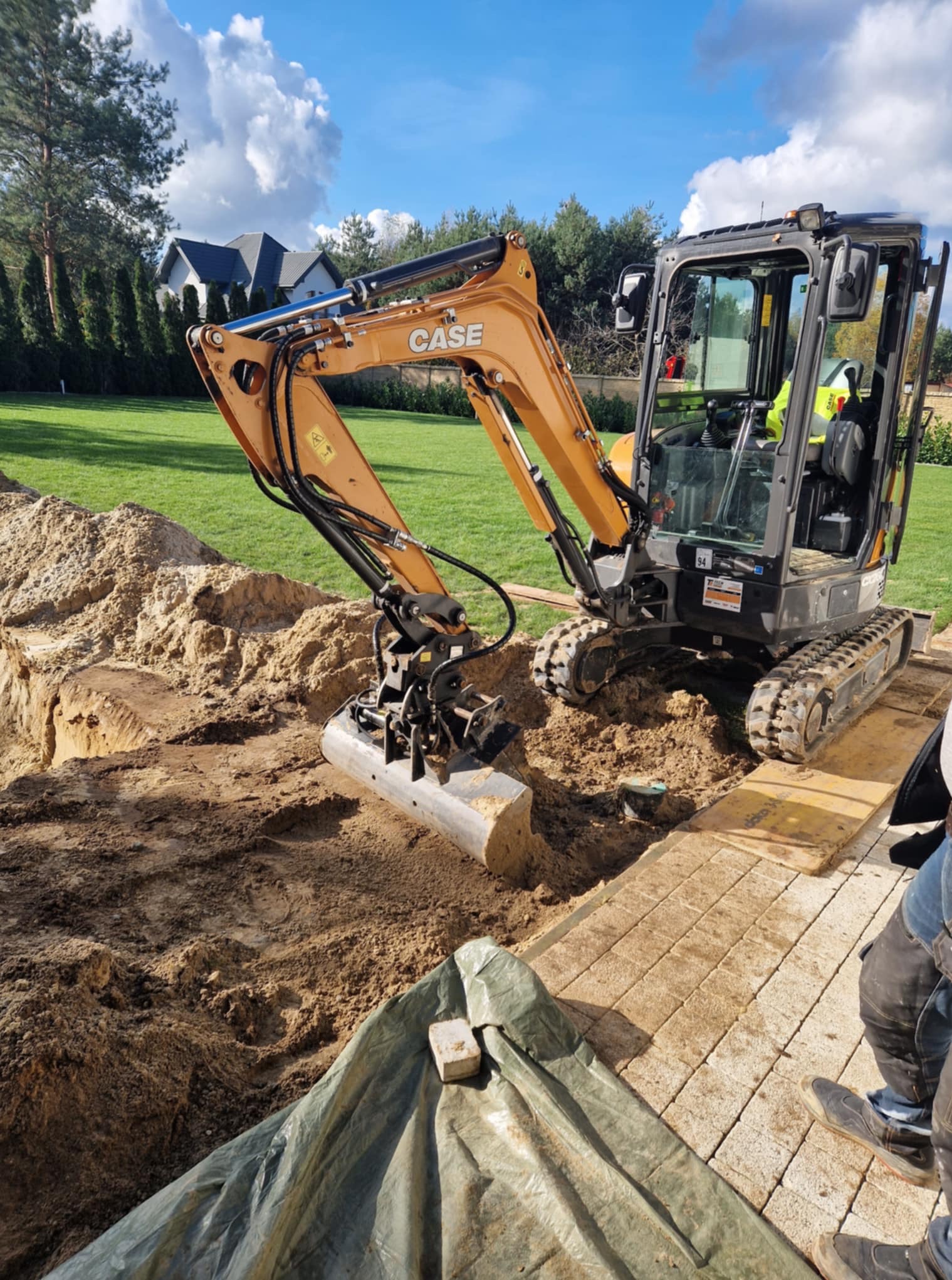 Żółto-czarna minikoparka Case w trakcie prac ziemnych na działce z trawą i kostką. Wykop pod instalacje lub fundamenty, widoczne drzewa i dom w tle.