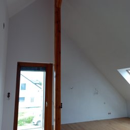 CraftPRO - Poddasze po remoncie: drewniana podłoga, świeżo pomalowane ściany, drewniany słup konstrukcyjny i okno z roletą. Widoczne punkty elektryczne na ścianie.