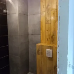 Nowoczesna łazienka w trakcie wykańczania: szare płytki wielkoformatowe i drewnopodobne panele, widoczne instalacje i fragment futryny w surowym stanie.