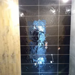 Błyszczące, czarne płytki z geometrycznym wzorem na ścianie, w trakcie montażu instalacji wodnej. Fragment ściany wyłożony płytkami imitującymi drewno i szare płytki.