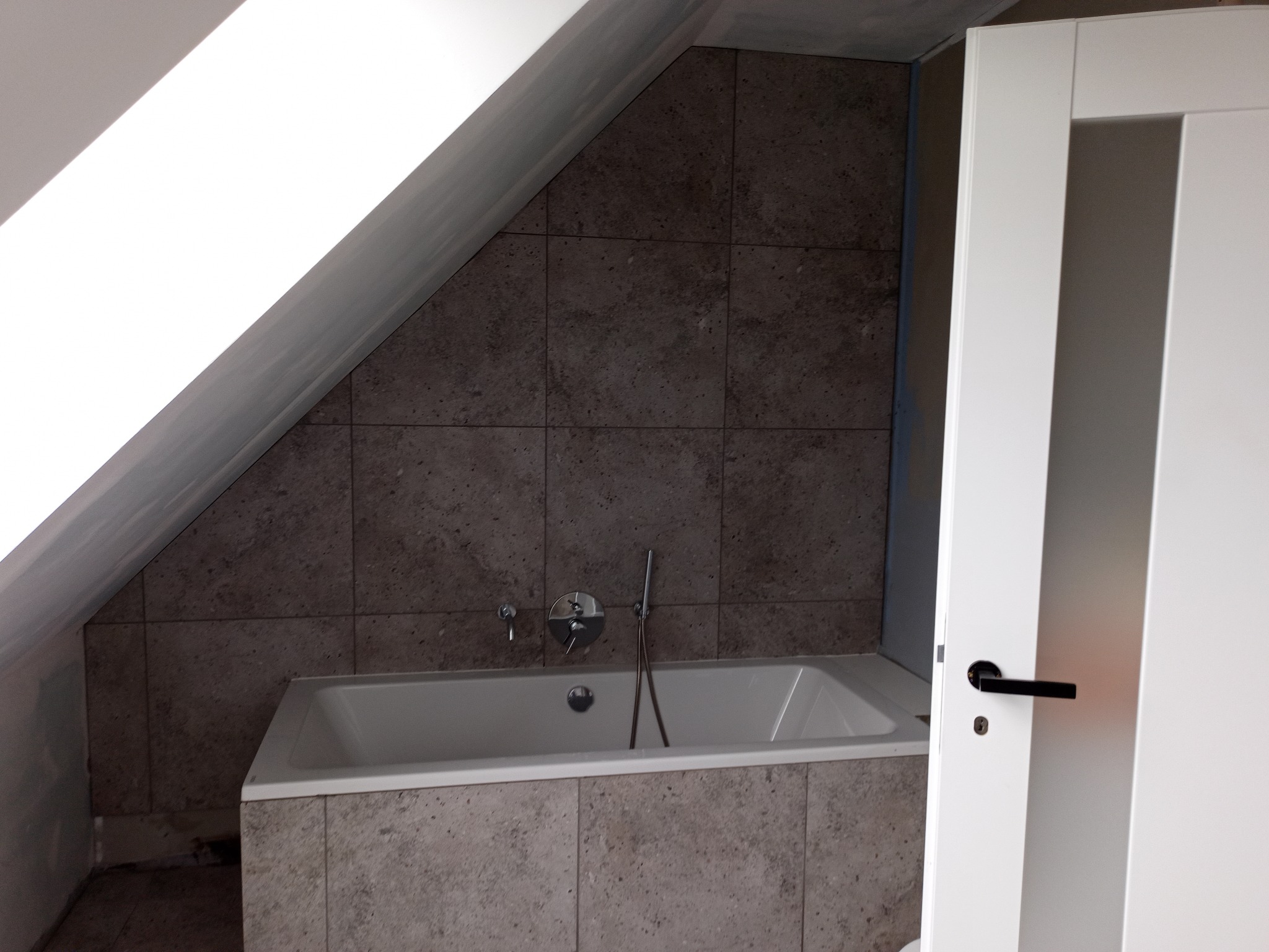 Wykończona łazienka na poddaszu: wanna obudowana płytkami, ściany w płytkach pod skosem dachu, białe drzwi z czarną klamką. Minimalistyczny design.