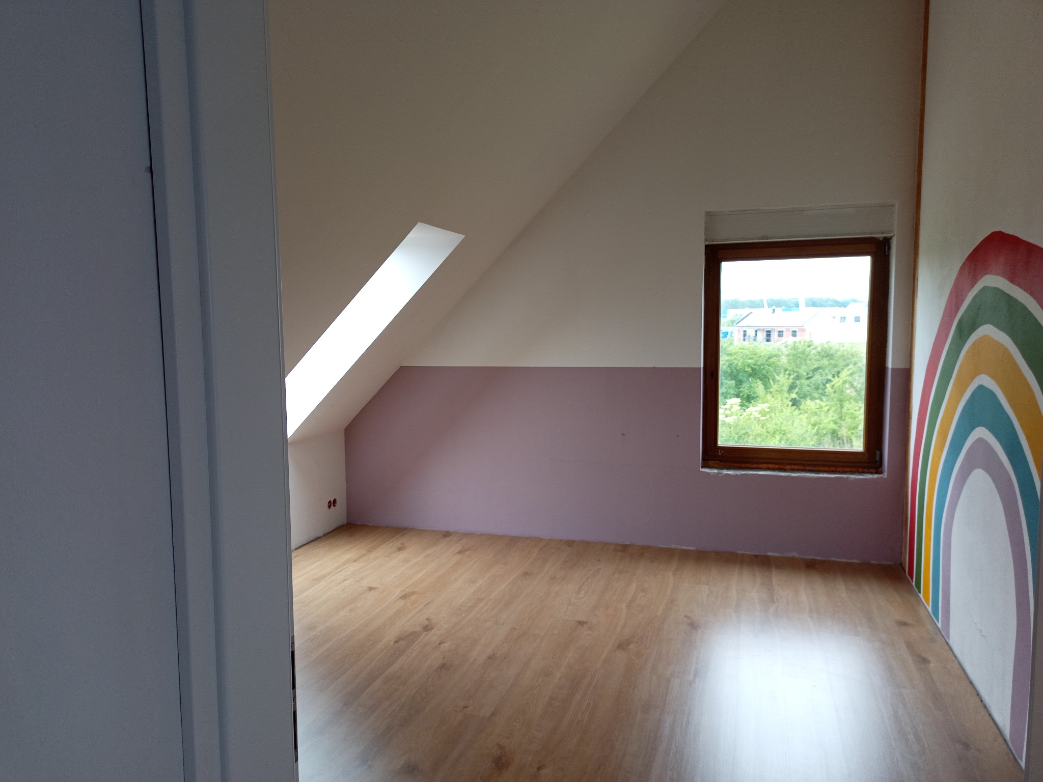 Wykończone poddasze z oknem dachowym i pomalowaną ścianą w tęczę w Wałbrzychu. Widoczna drewniana podłoga i biało-różowe ściany. Częściowo otwarte białe drzwi.