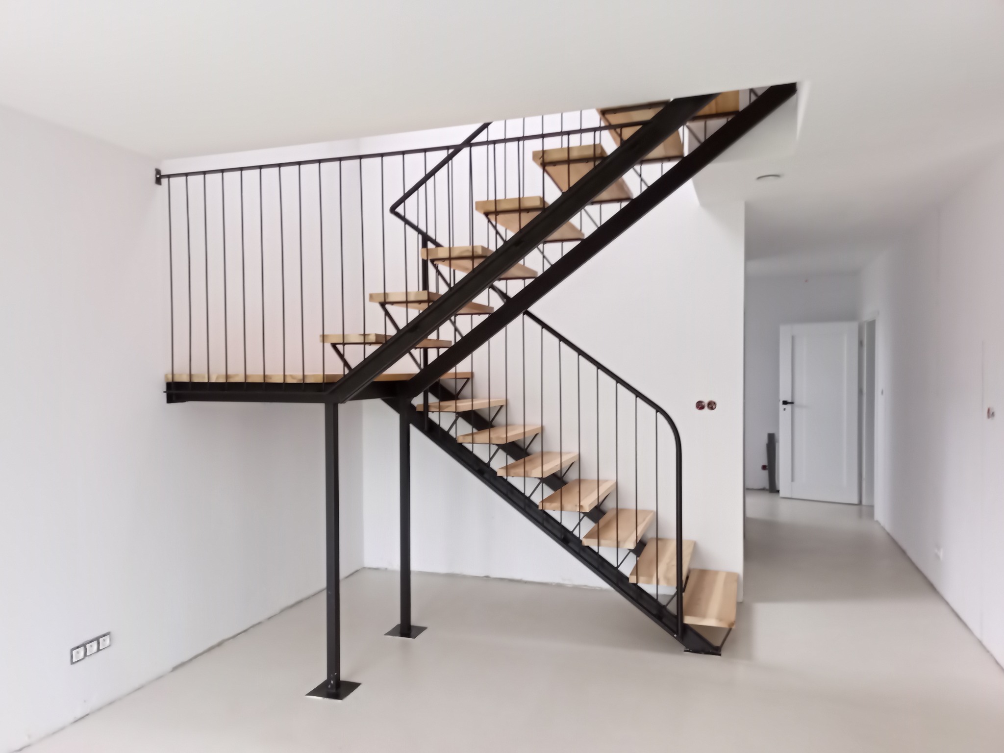Nowoczesne schody z drewnianymi stopniami i czarną metalową balustradą w jasnym, minimalistycznym wnętrzu. Widoczne gniazdka elektryczne na ścianie i drzwi w tle.