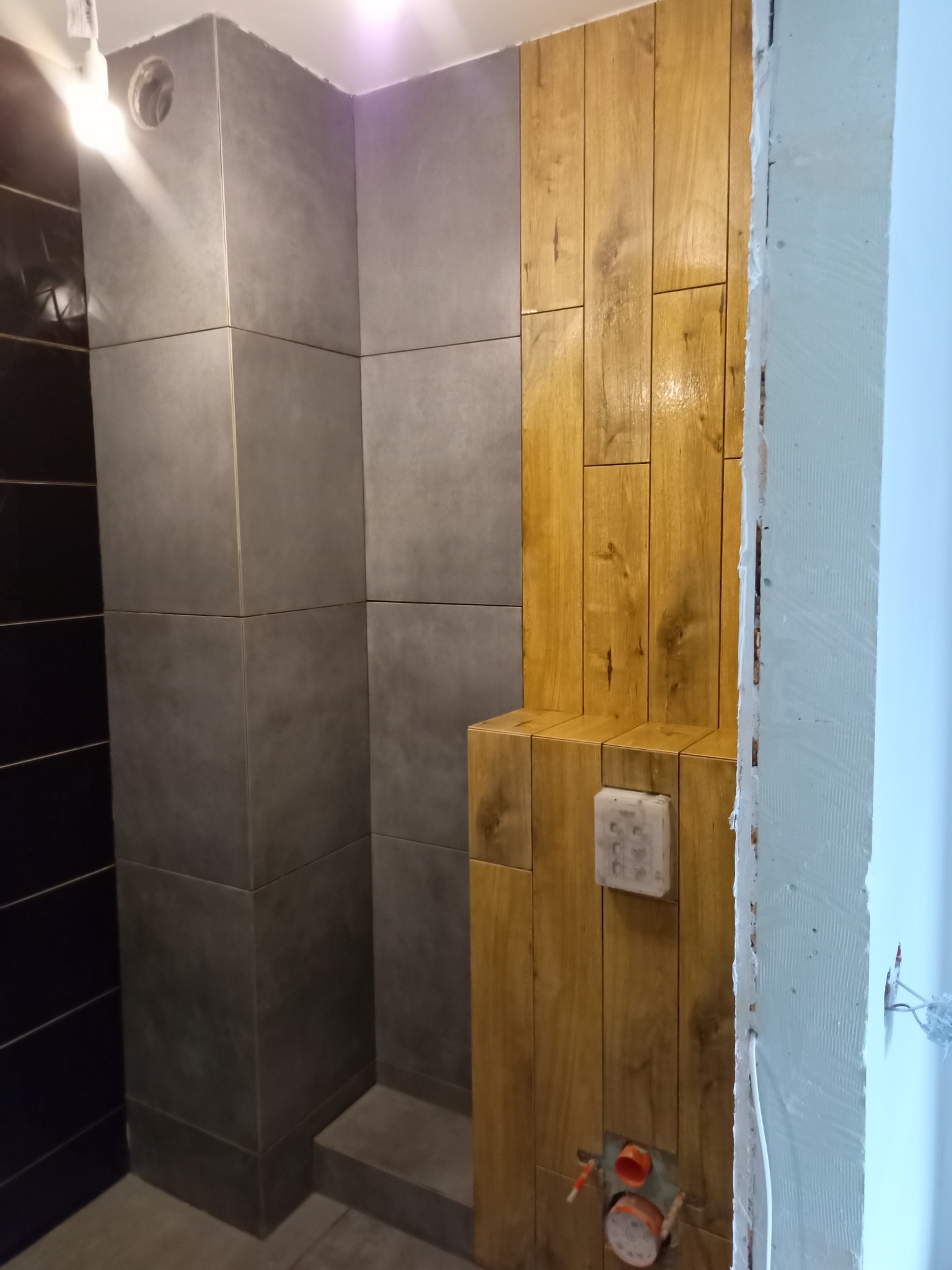 Łazienka w trakcie wykończenia: szare, kwadratowe płytki ceramiczne kontrastują z drewnopodobnymi panelami. Widoczne instalacje i fragment ściany w stanie surowym.