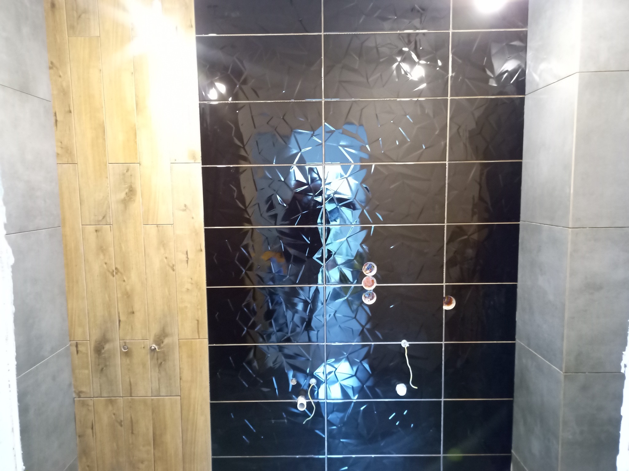 Częściowo wykończona łazienka z czarnymi, błyszczącymi płytkami o trójwymiarowej fakturze, drewnopodobnymi płytkami i szarymi płytkami na ścianie.