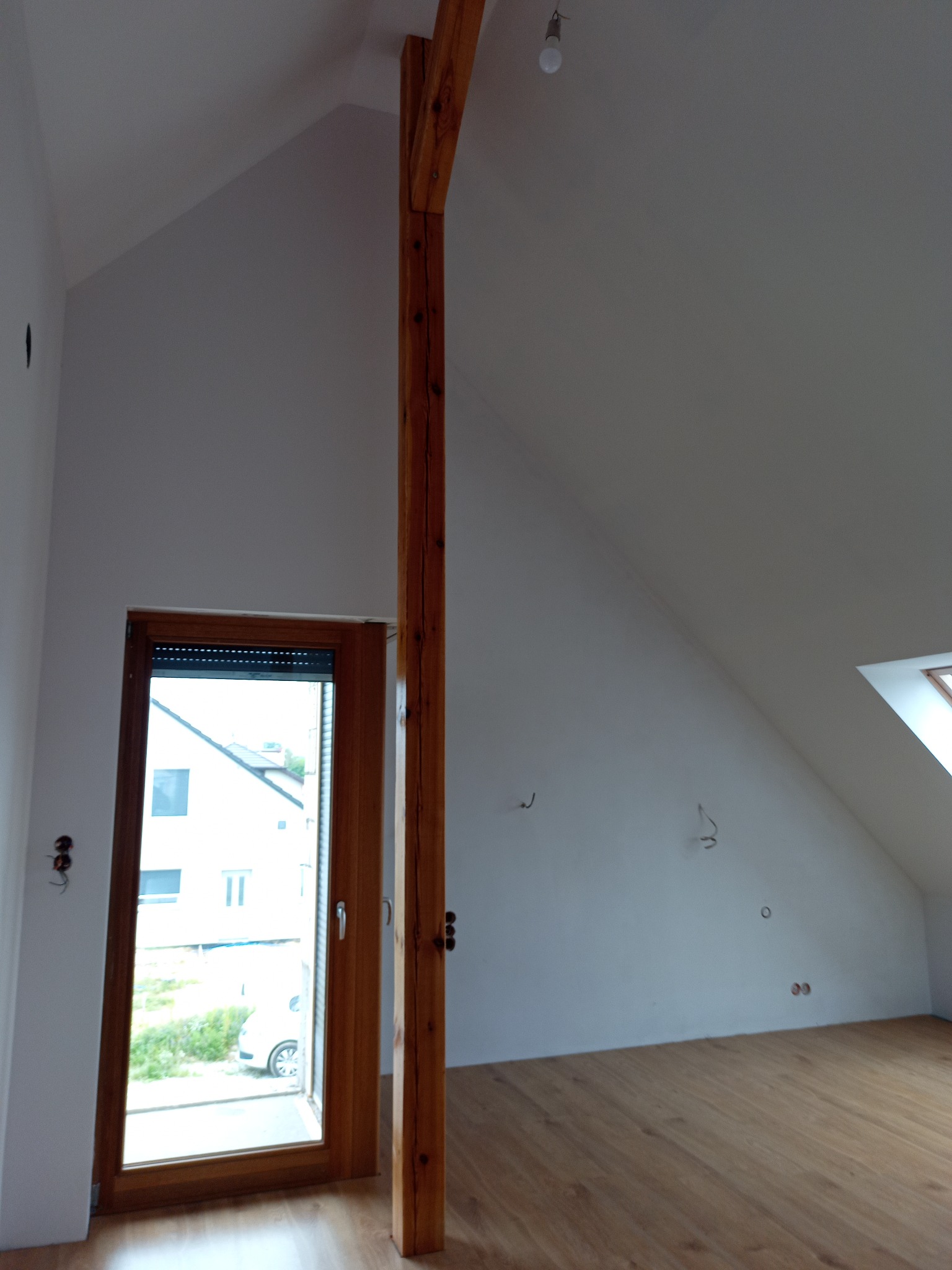 Poddasze po remoncie: drewniana podłoga, okno z roletą, drewniany słup konstrukcyjny. Ściany pomalowane na biało, widoczne punkty elektryczne.