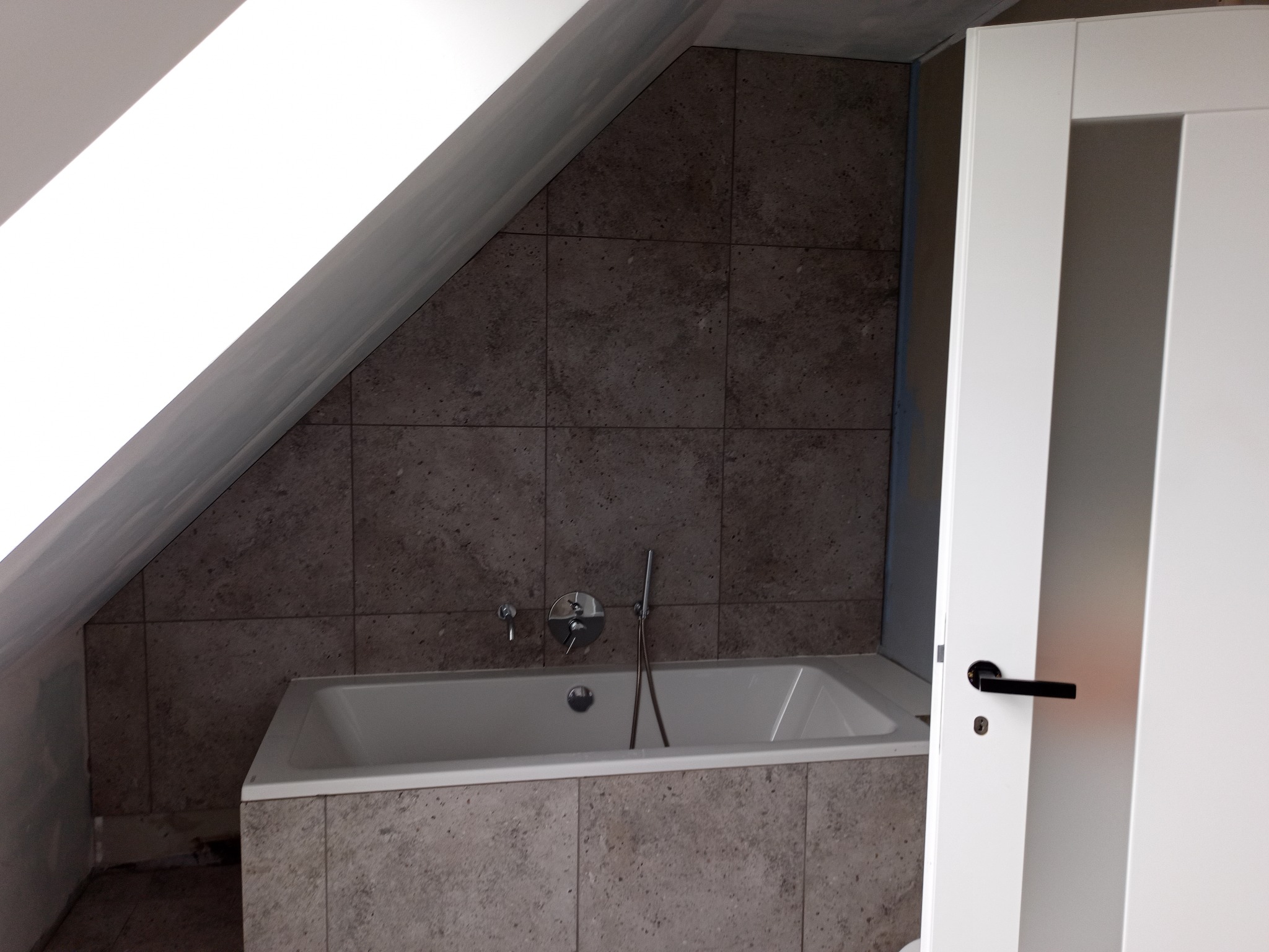 Wykończona łazienka na poddaszu: wanna obudowana płytkami, ściany wyłożone płytkami w odcieniach szarości, skośny sufit, białe drzwi z przeszkleniami.