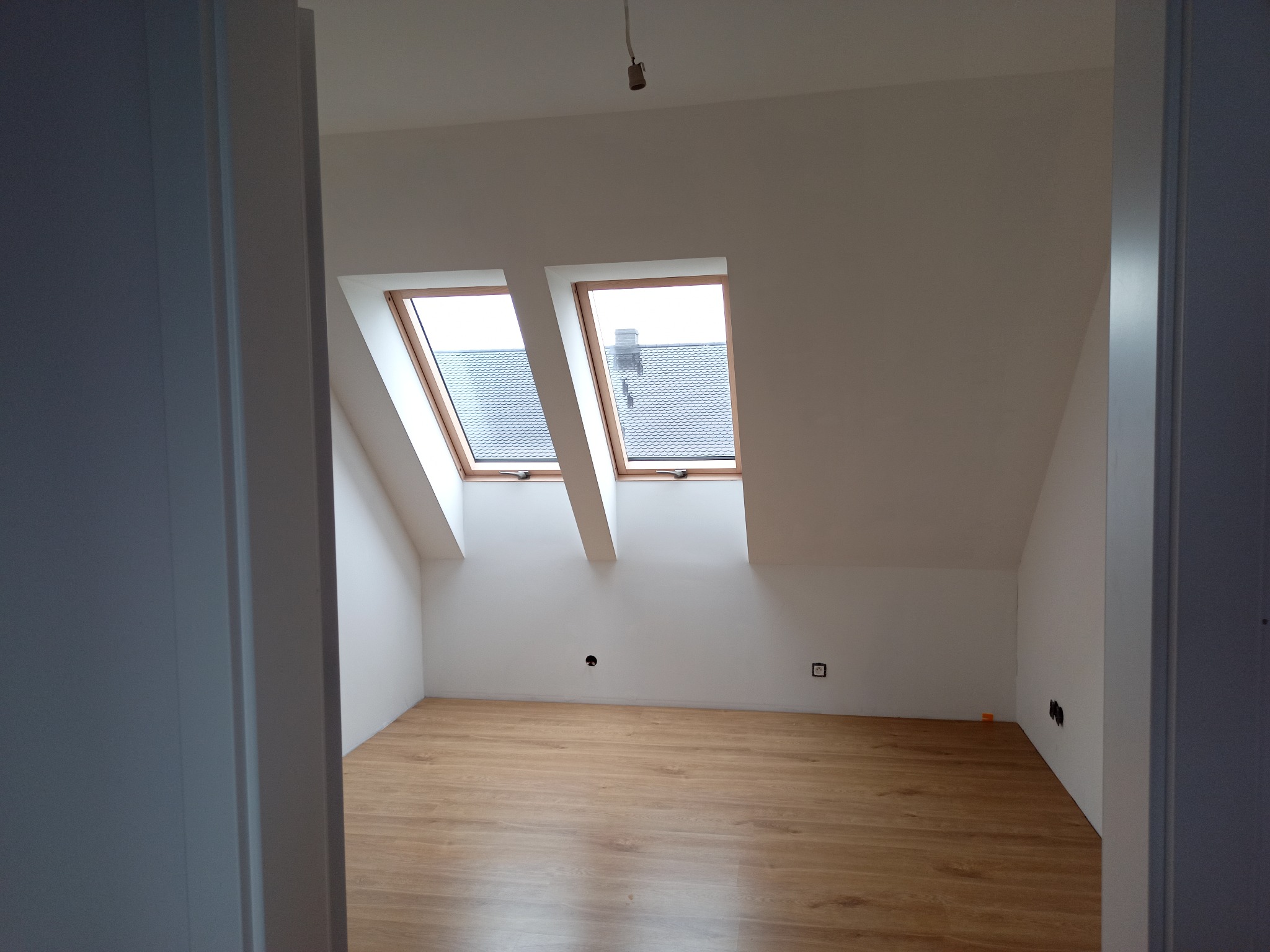 Poddasze z drewnianą podłogą i dwoma oknami dachowymi, widoczne przez otwarte drzwi. Ściany białe, gniazdka elektryczne zamontowane. Minimalistyczny wystrój.