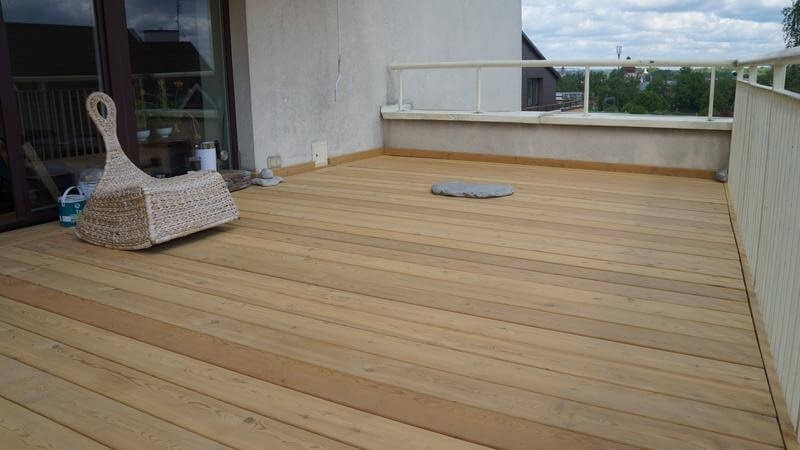 Jasny taras drewniany z krzesłem z rattanu i widokiem na zieleń. Deski ułożone równolegle, balustrada biała. Kamień dekoracyjny na podłodze.