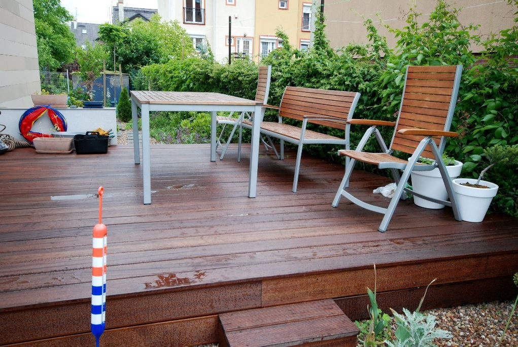 Drewniany taras z meblami ogrodowymi: stół z szarymi nogami, ławka i krzesło na tle zieleni. Na pierwszym planie zabawka w biało-czerwono-niebieskie paski.