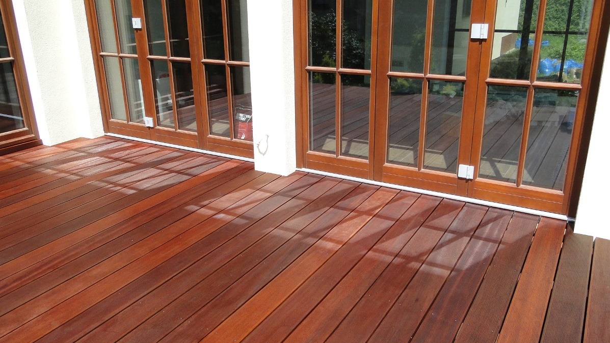Wykończony taras drewniany z egzotycznego drewna, z widocznymi promieniami słońca i eleganckimi, przeszklonymi drzwiami tarasowymi w tle.
