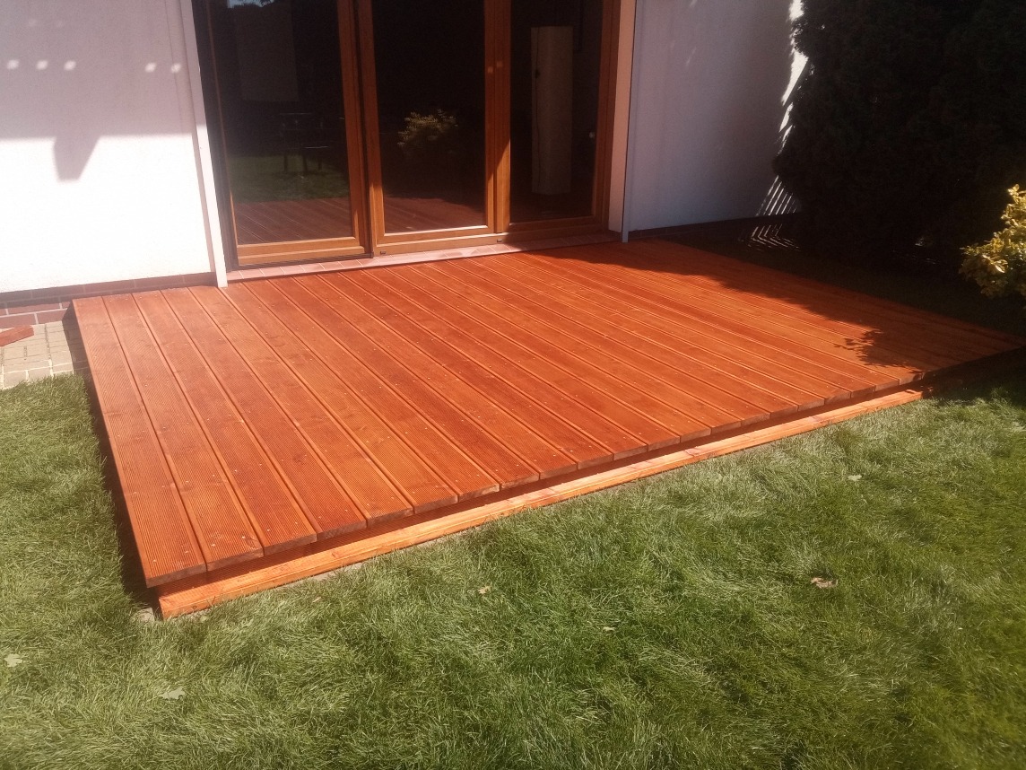 Nowoczesny taras drewniany z desek w ciepłym odcieniu brązu, przylegający do białego budynku z przeszklonymi drzwiami, otoczony zielonym trawnikiem.