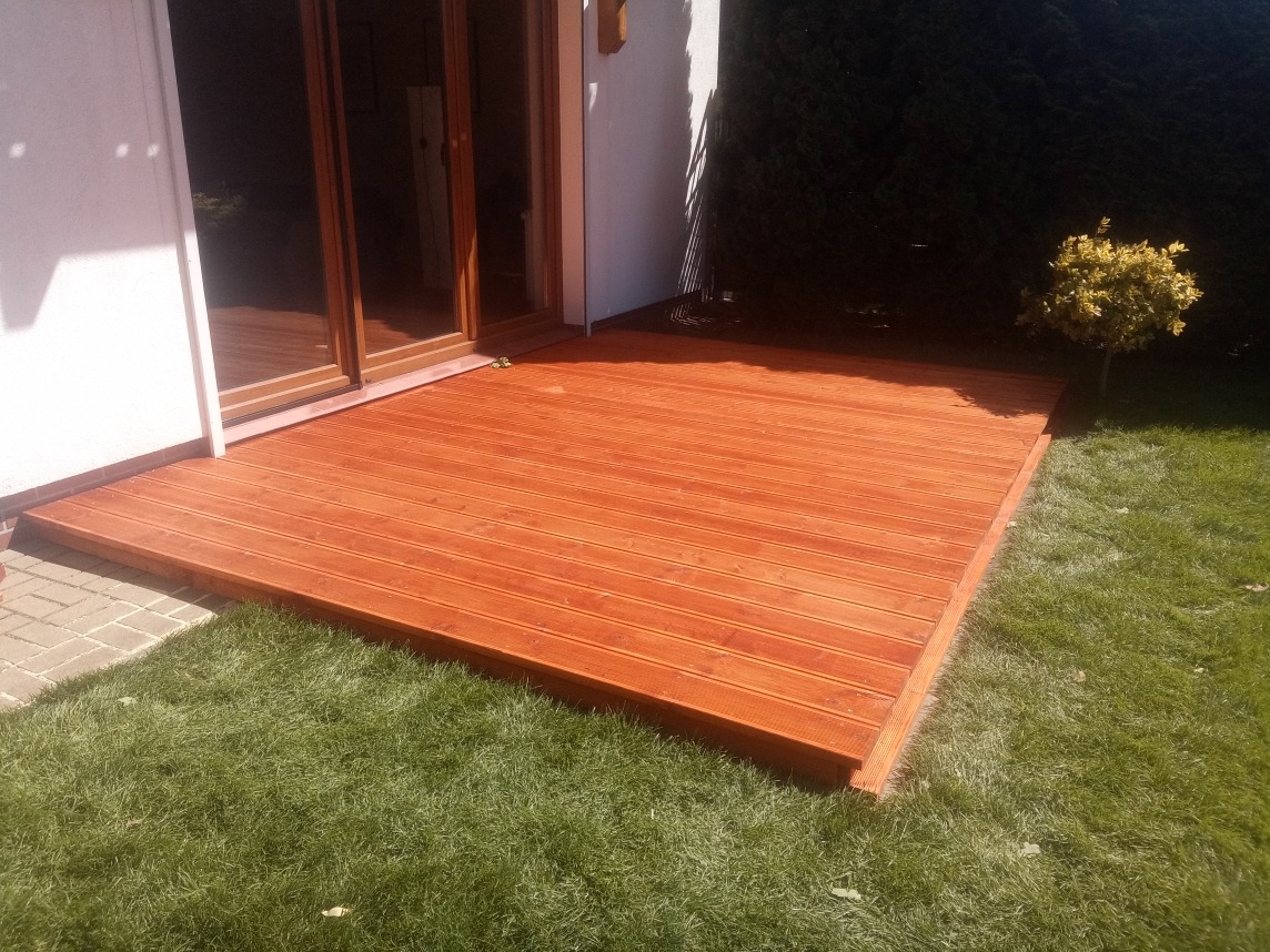 Ukończony taras drewniany z desek w ciepłym odcieniu brązu, przylegający do domu z widoczną zielenią trawnika i krzewu w tle, w słoneczny dzień.