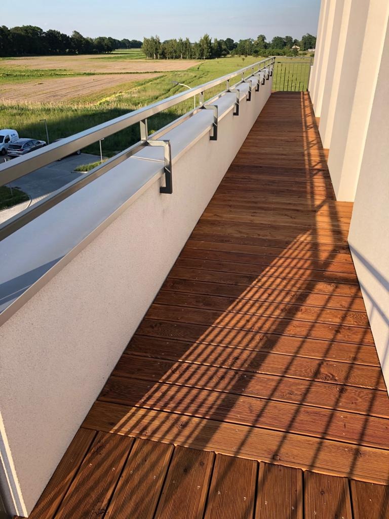 Długi balkon z drewnianą podłogą, metalową balustradą i widokiem na zielone pola w oddali, oświetlony popołudniowym słońcem rzucającym cienie.