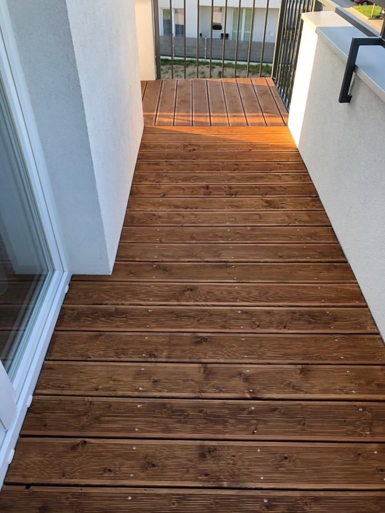 Nowoczesny taras z desek kompozytowych w ciepłym odcieniu brązu, widok z góry. Deski ułożone równolegle, widoczne wkręty. Białe ściany po bokach.