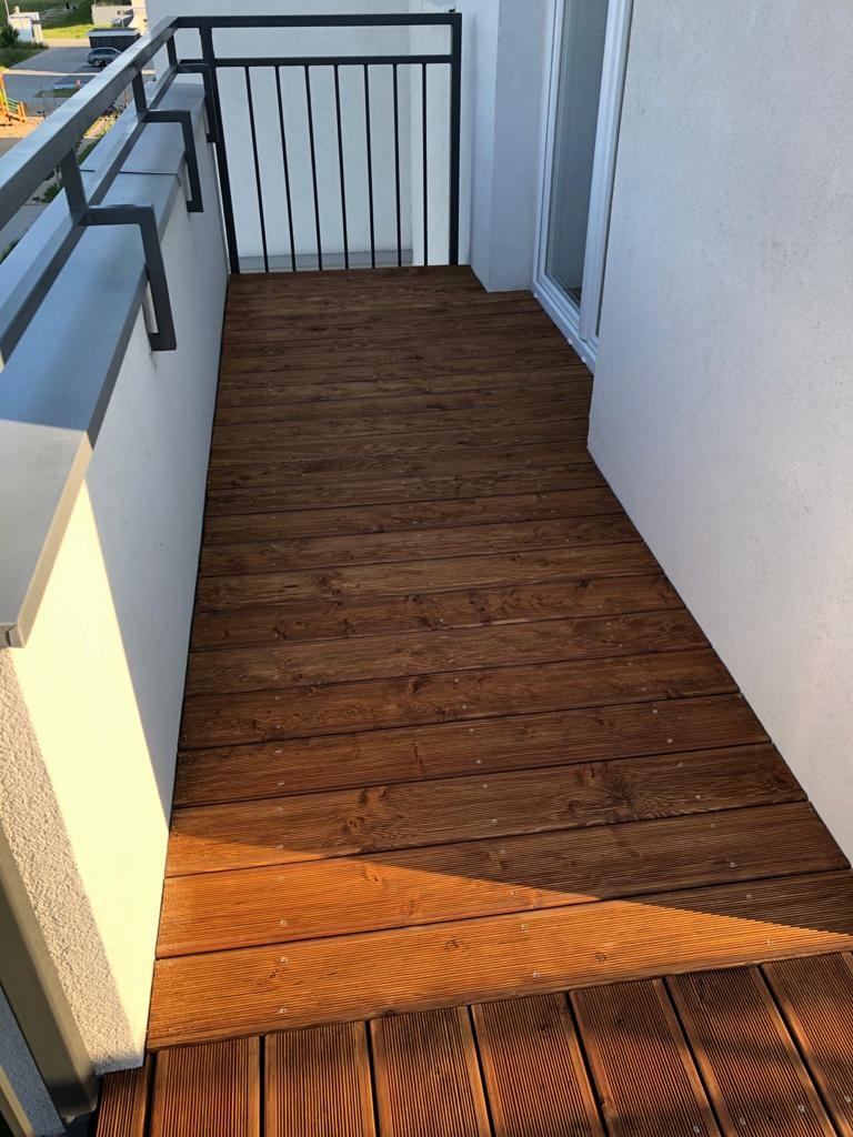 Balkon z drewnianą podłogą z desek o ciepłym brązowym odcieniu, z metalową barierką i białymi ścianami. Widok z góry, oświetlony naturalnym światłem.