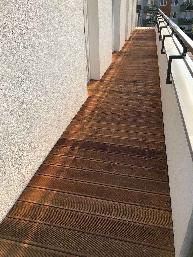 Długi balkon z ciemnobrązową deską podłogową, jasną ścianą i metalową balustradą. Widok wzdłuż balkonu, w oddali budynek mieszkalny.