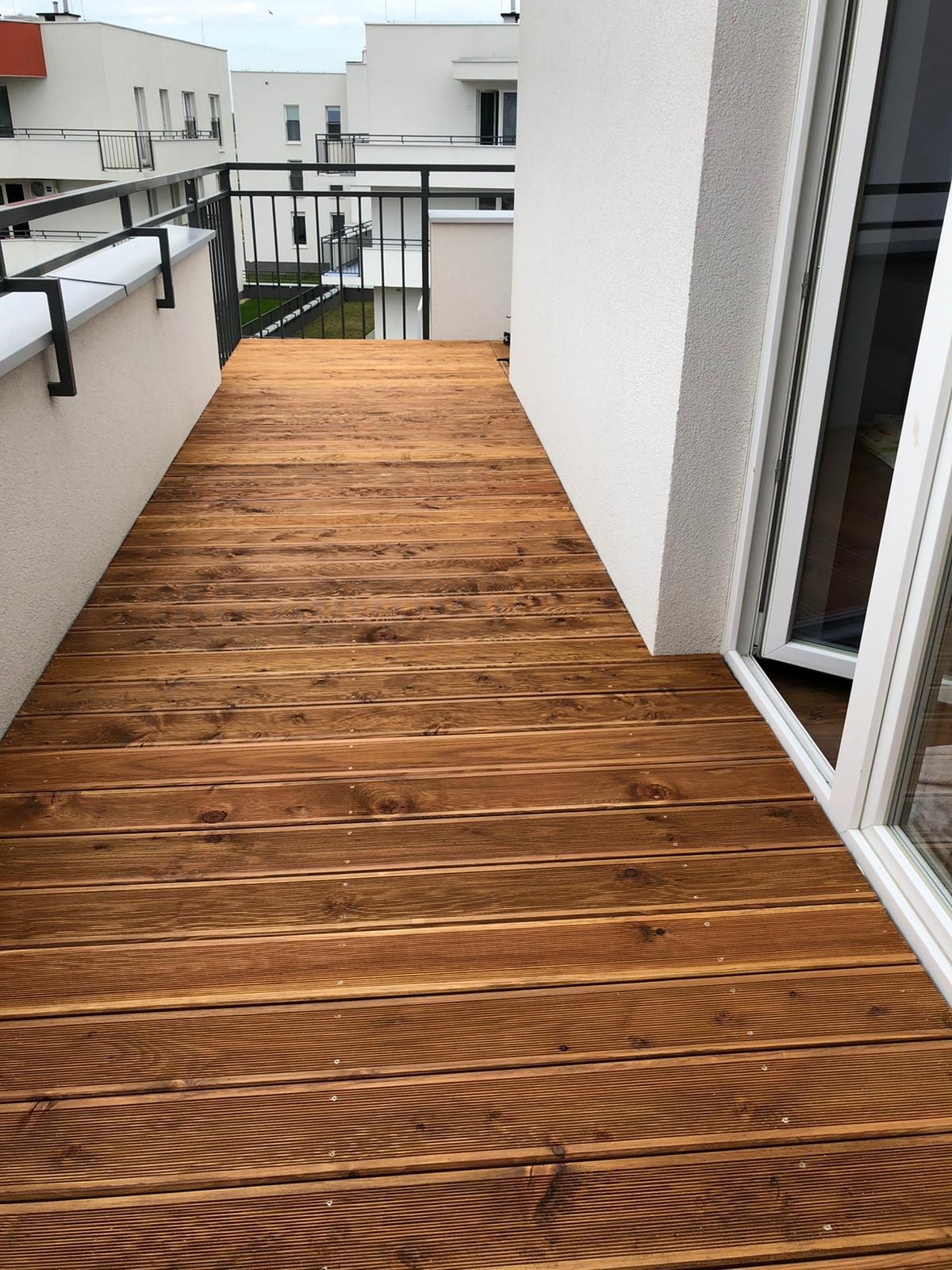 Nowoczesny taras drewniany na balkonie z deskami w ciepłym odcieniu brązu, widok z góry, białe ściany i czarne barierki. Estetyczne wykończenie.