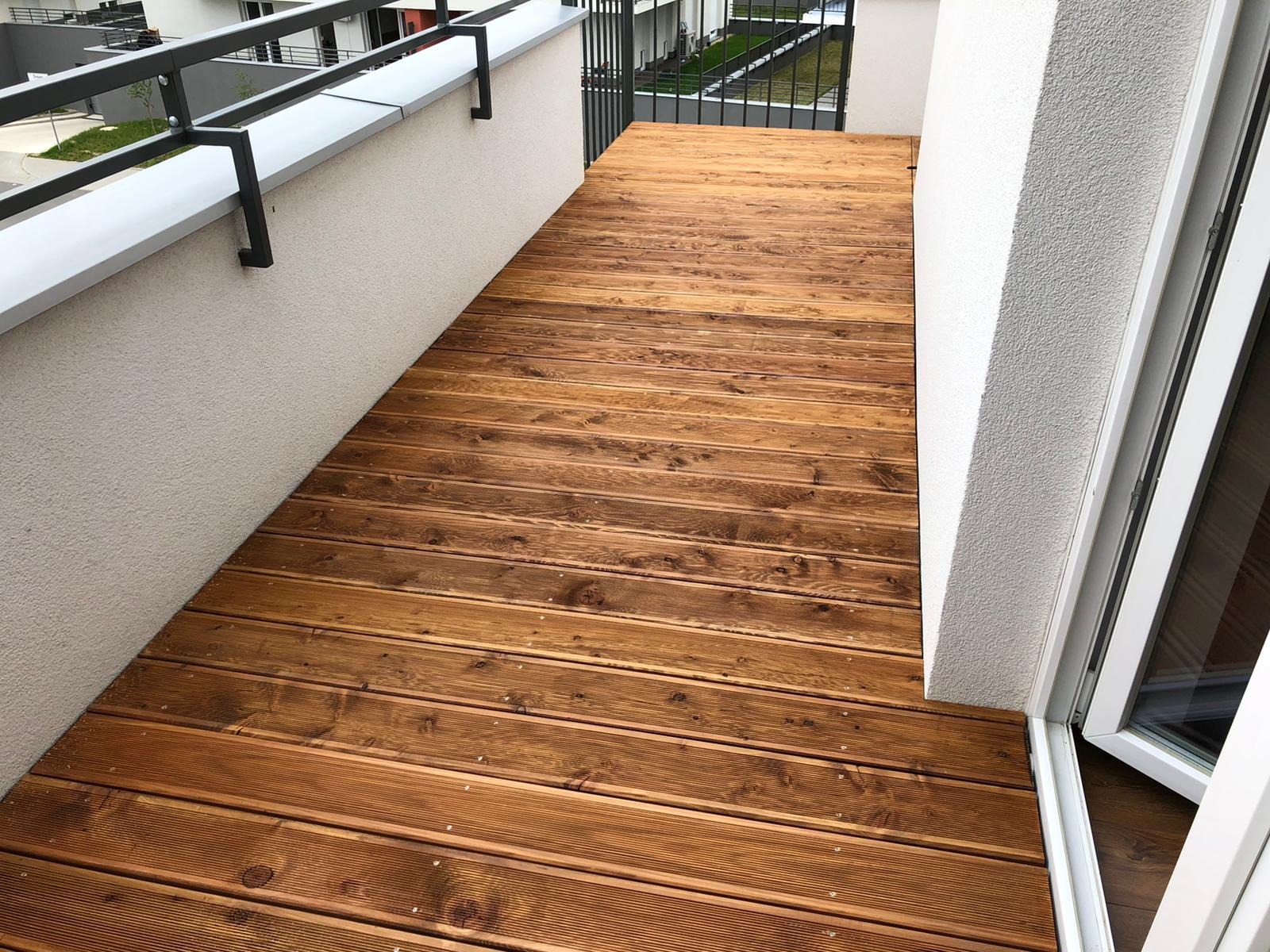 Nowoczesny balkon z drewnianą podłogą, widok z góry. Deski ułożone równolegle, w tle biała ściana i balustrada. Częściowo otwarte drzwi balkonowe.