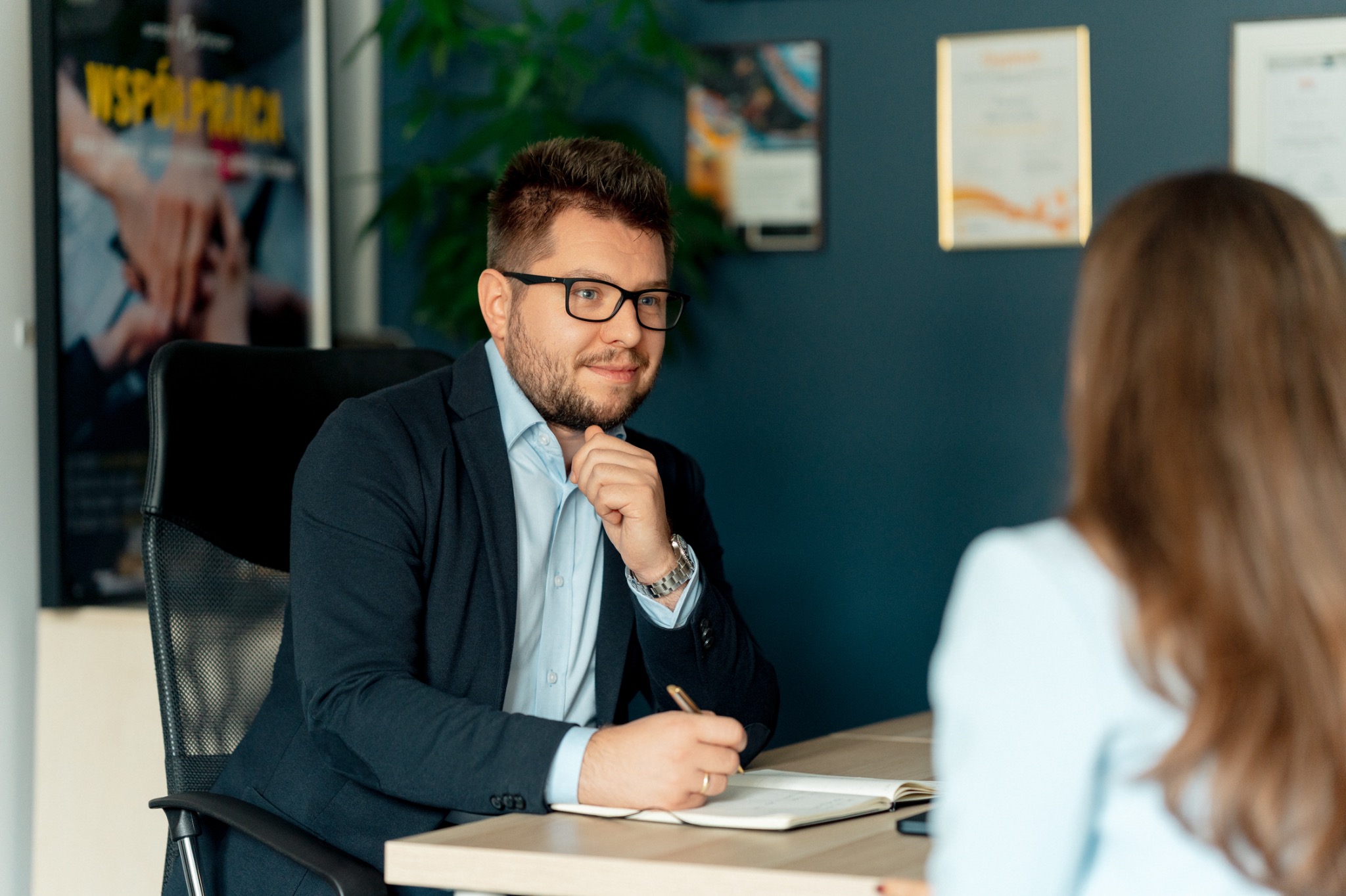 Spotkanie doradcze: mężczyzna w garniturze notuje w notatniku podczas rozmowy z klientką w biurze z dyplomami na ścianie i rośliną w tle.