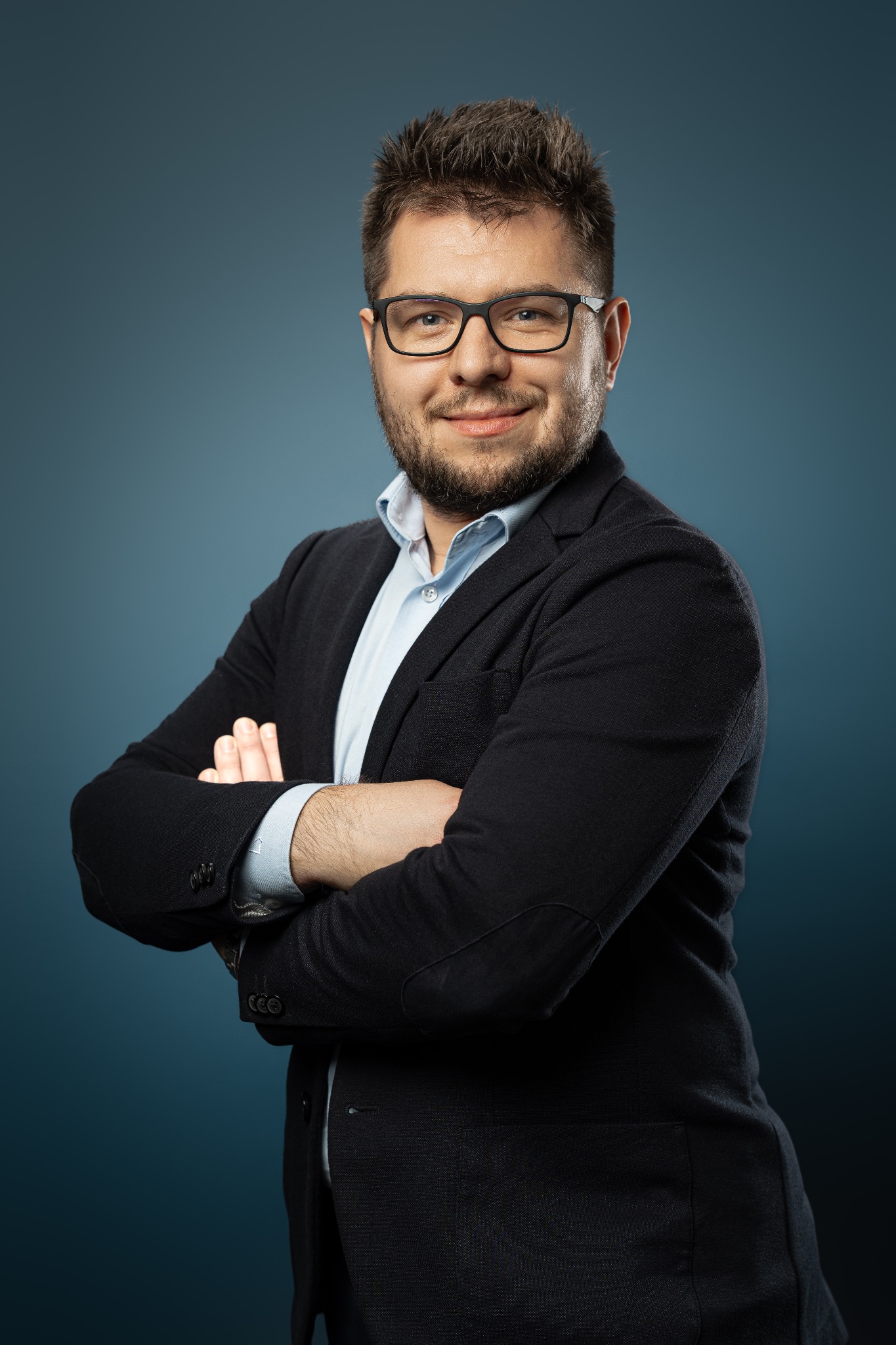 Portret mężczyzny w okularach, ubrany w ciemną marynarkę i jasną koszulę, z założonymi rękami na tle ciemnoniebieskiego gradientu. Uśmiechnięty, profesjonalny wizerunek.