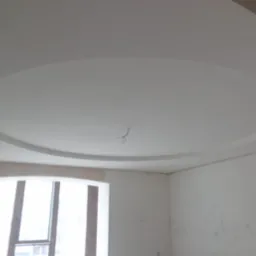 Biały sufit z okrągłą dekoracją gipsową, widoczne wystające przewody, ściany przygotowane do malowania, okno z ciemnymi ramami.