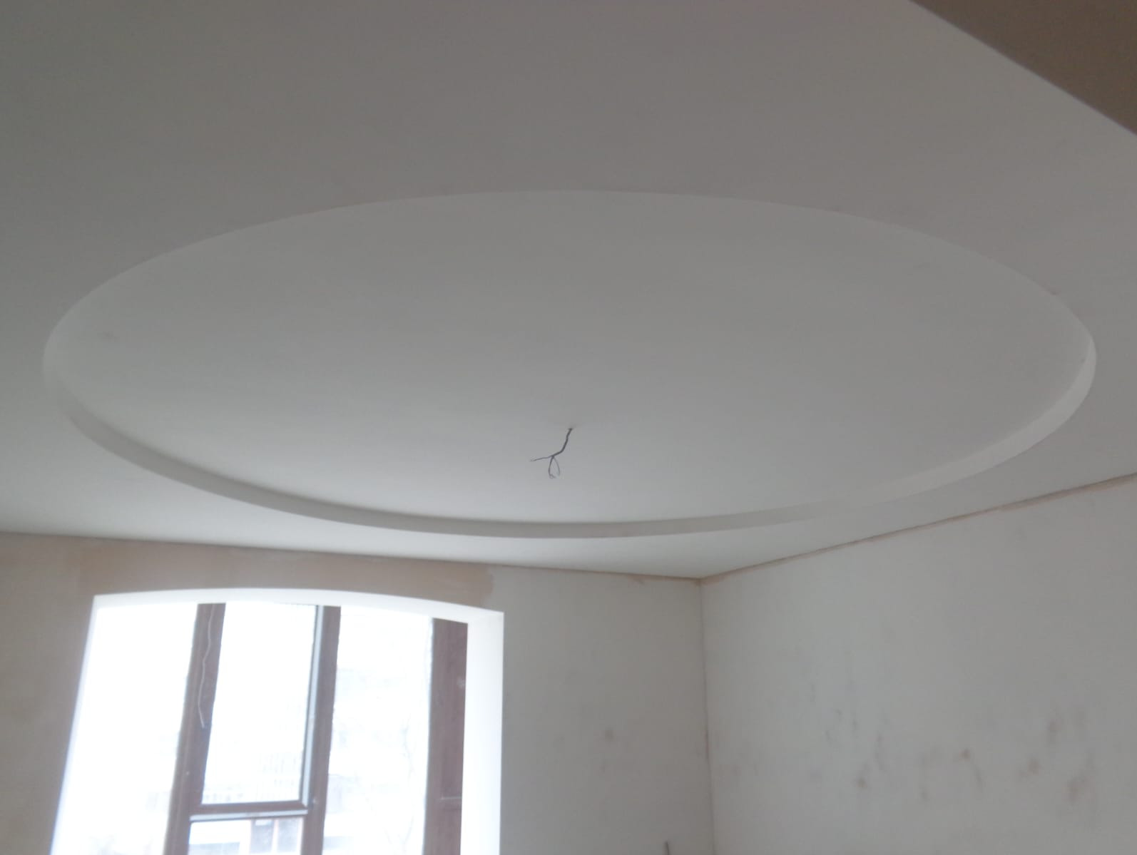 Biały sufit z okrągłą dekoracją gipsową, widoczne wystające przewody, ściany przygotowane do malowania, okno z ciemnymi ramami.