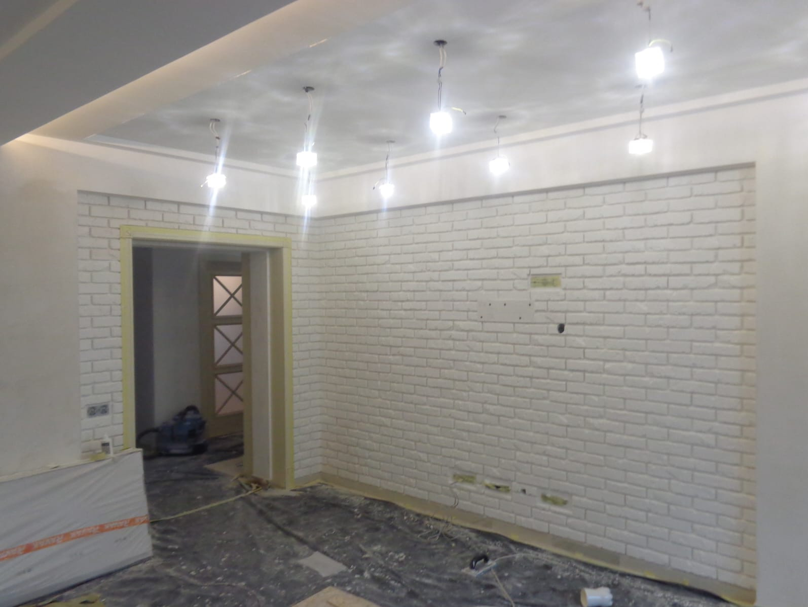 Biała ceglana ściana z oświetleniem sufitowym w odnowionym wnętrzu. Widoczne przygotowania do dalszych prac wykończeniowych i zabezpieczona podłoga folią.