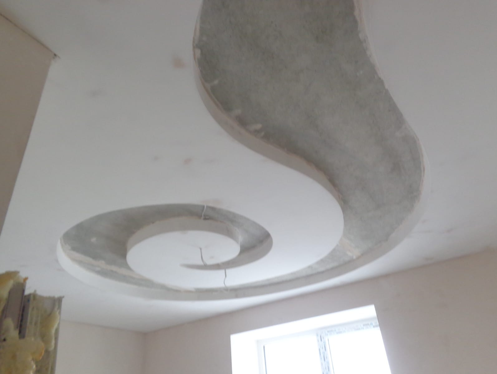 Dekoracyjny sufit podwieszany z płyty gipsowej w kształcie spirali, częściowo wykończony gładzią, widok z dołu, surowe wnętrze z oknem.