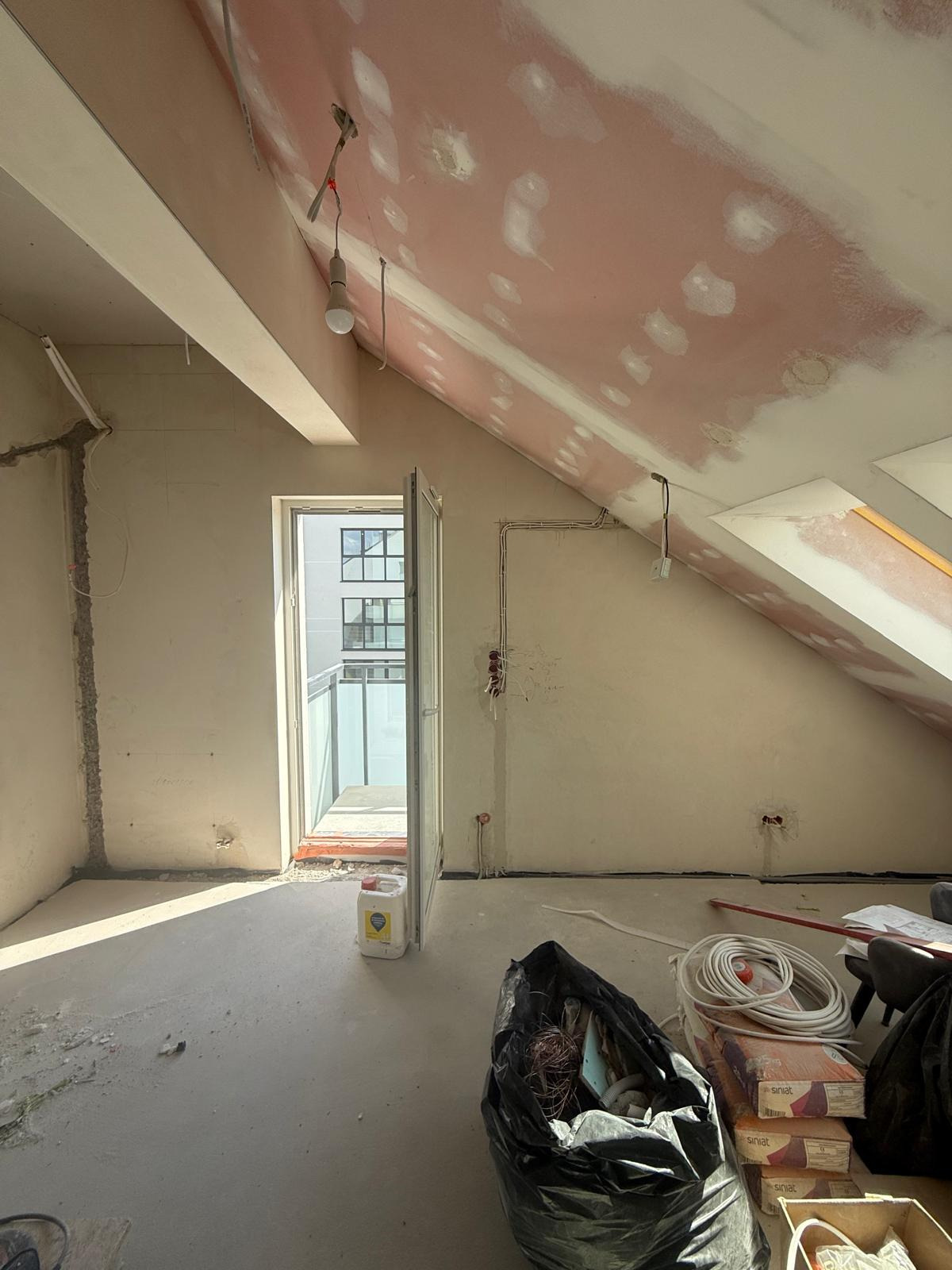 Poddasze w trakcie remontu: ściany po gładzi, sufit z widocznymi punktami po łatach, otwarte drzwi balkonowe, materiały budowlane na podłodze.